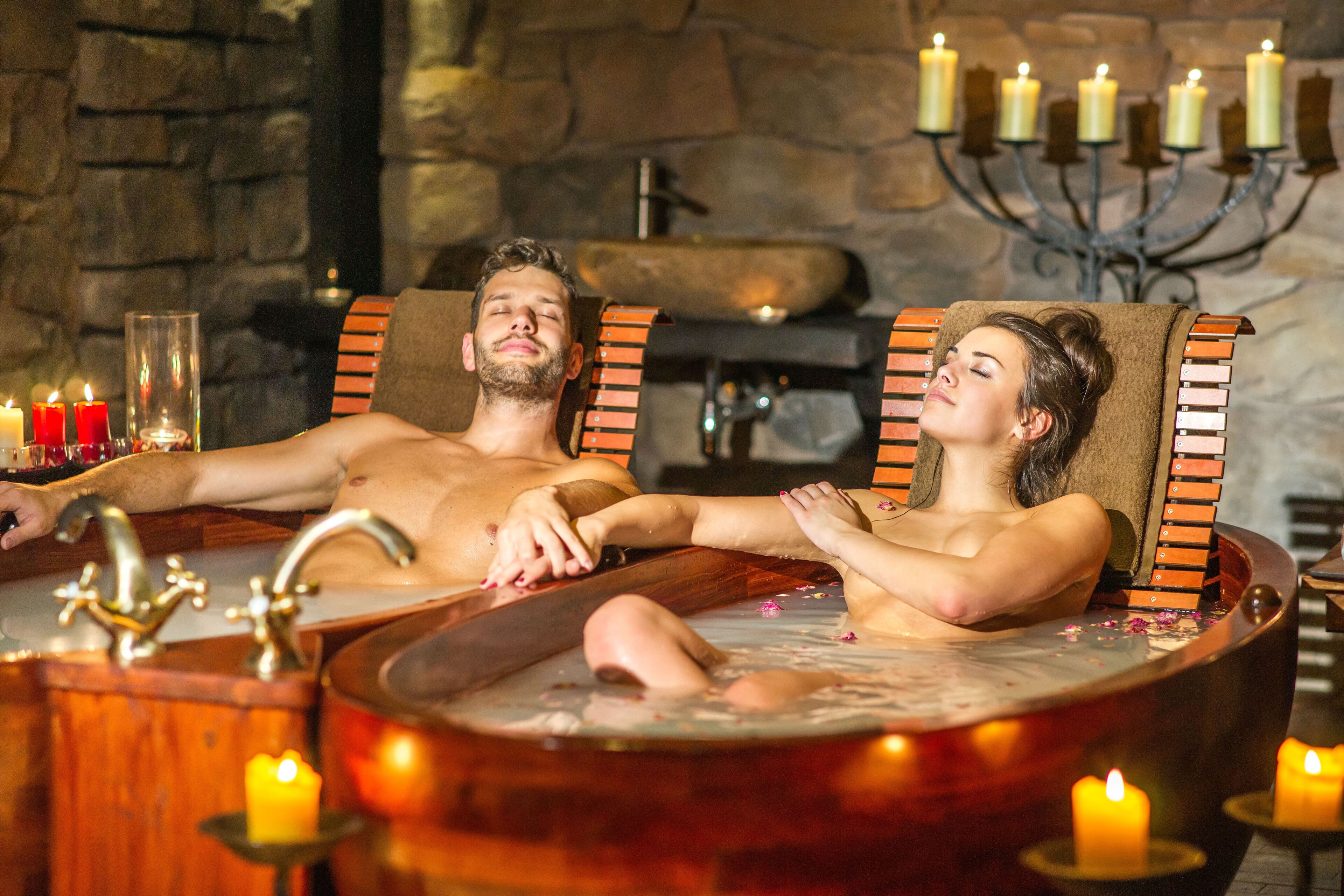 A couple relaxing in a wooden bathtub with candles and flowers around them.