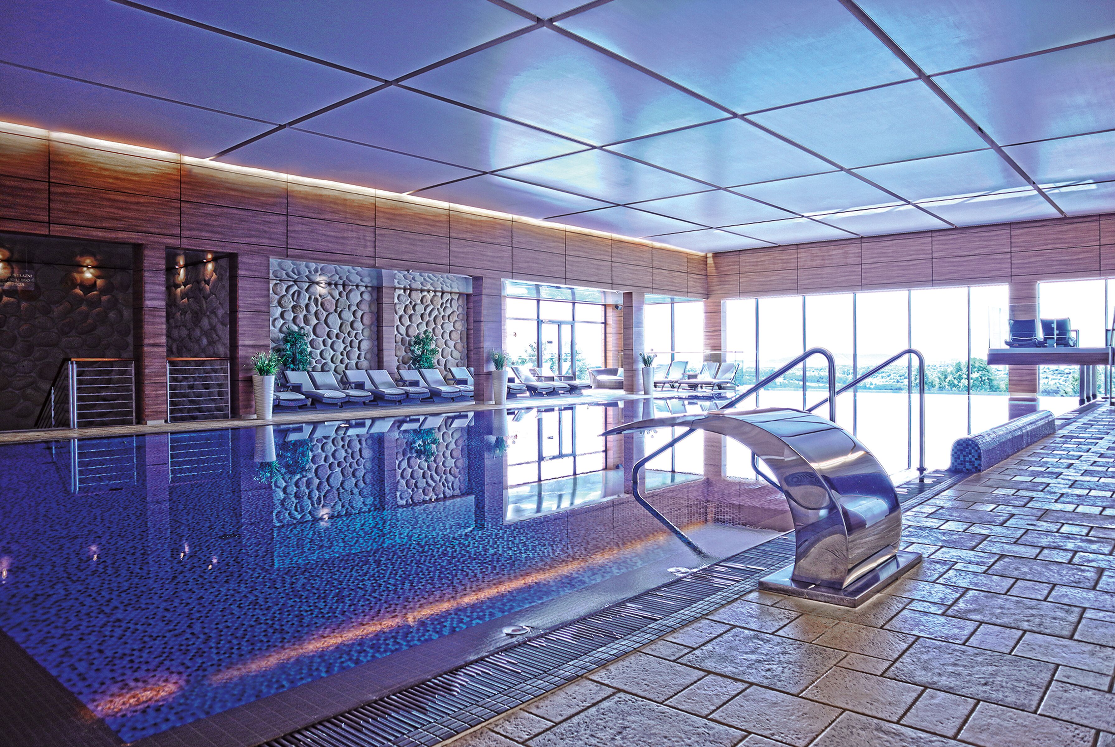 An indoor swimming pool with a clear view of the pool, lounge chairs, and a staircase leading to an upper level.