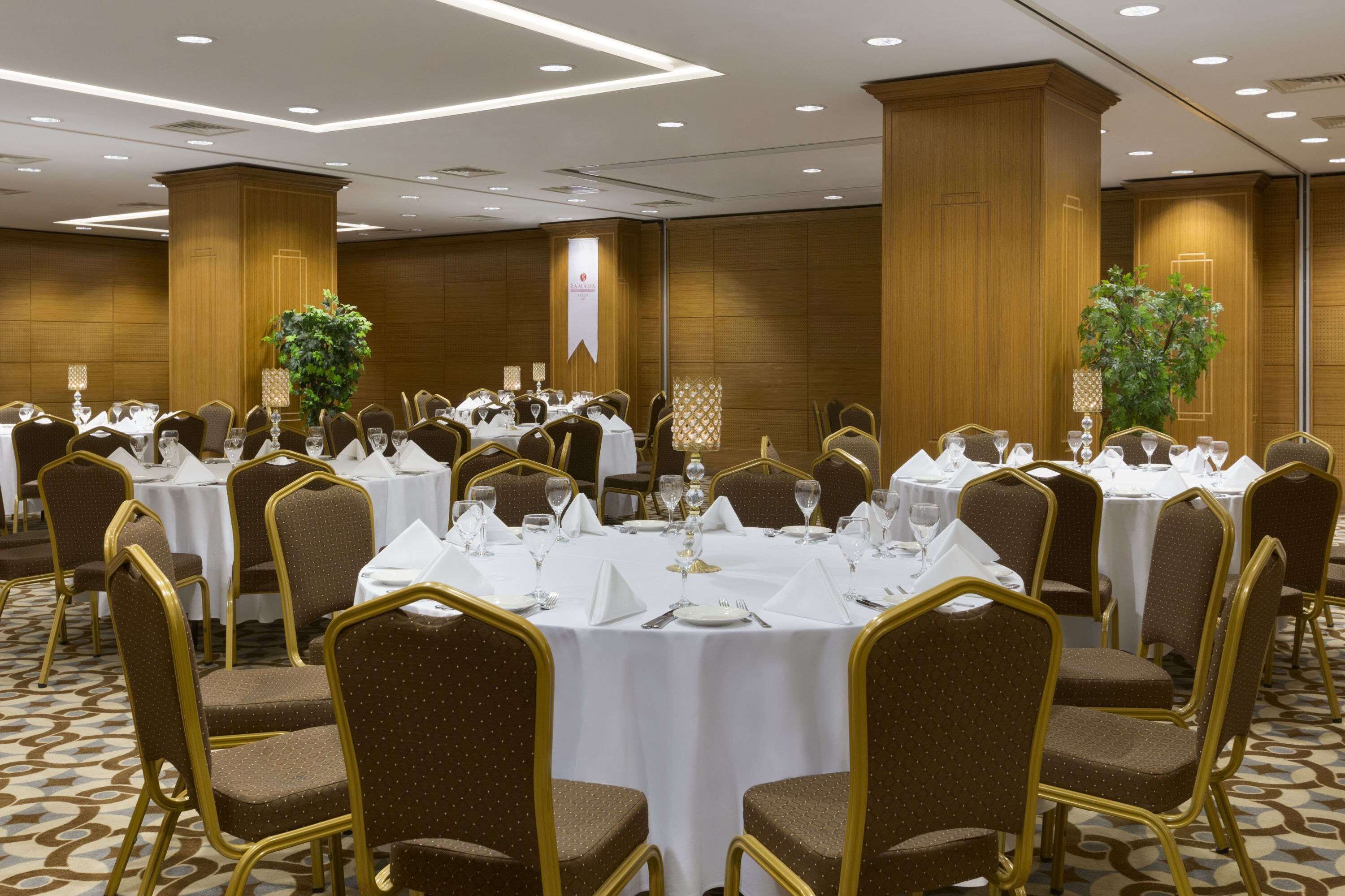 Un salón de banquetes con mesas redondas dispuestas para comer, rodeado de sillas tapizadas con detalles dorados.