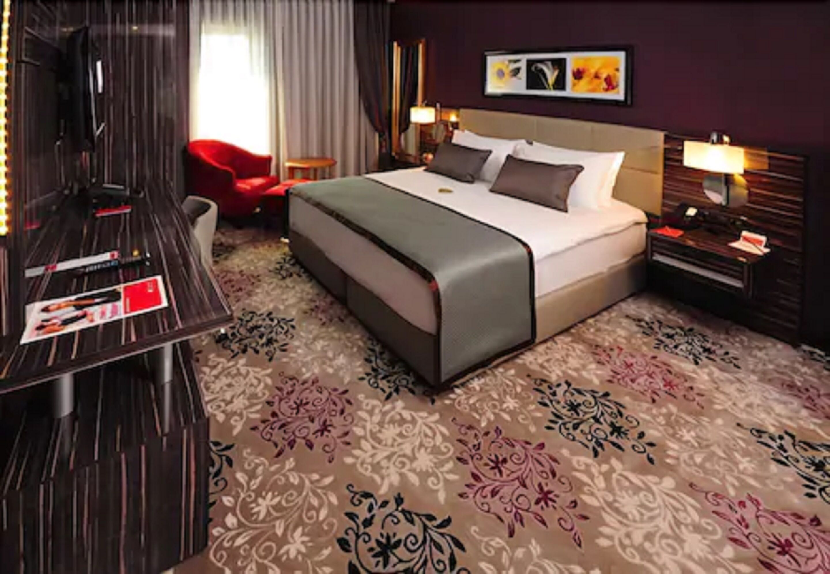 A hotel room with a large bed, a desk, a red chair, and a patterned carpet.