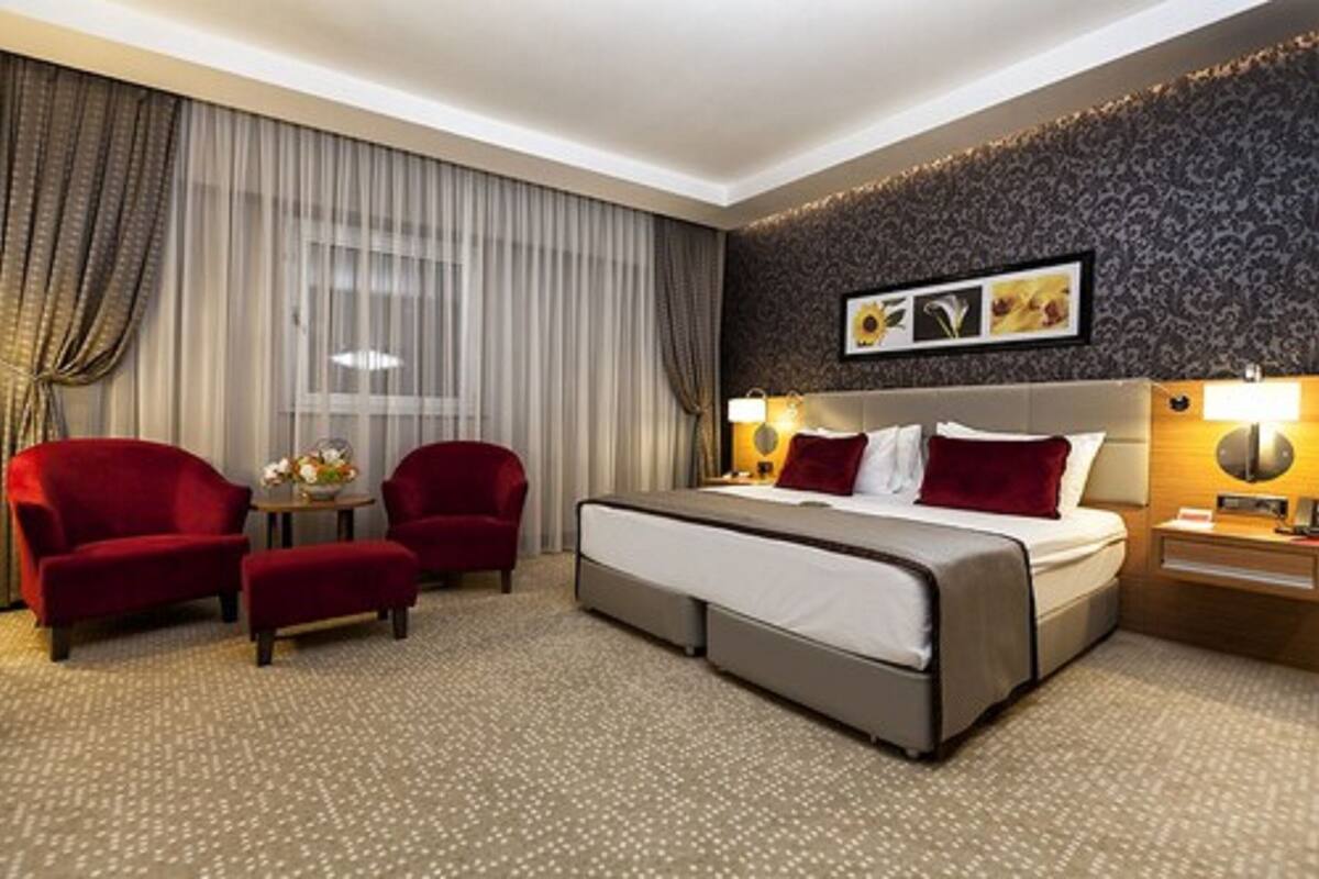 Habitación de hotel con una cama grande, dos sillones rojos, una mesita con un florero y un ventanal con cortinas.