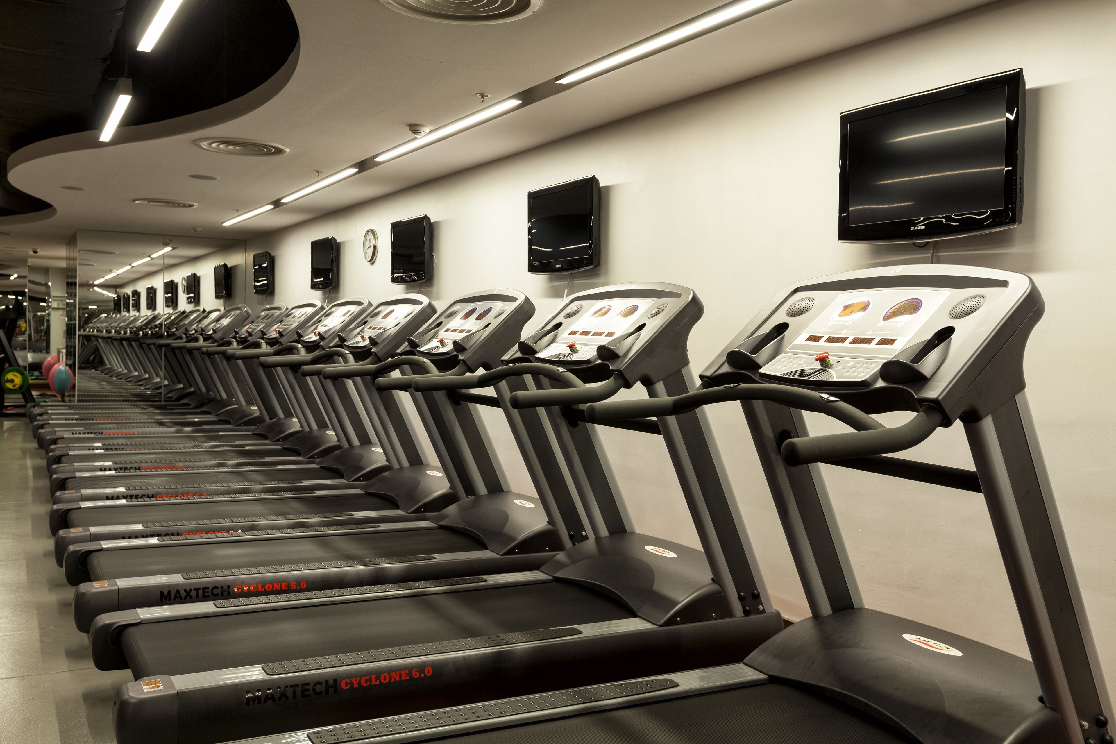 Una fila de cintas de correr en un gimnasio con piso plano, un televisor montado en la pared y un reloj en la pared.
