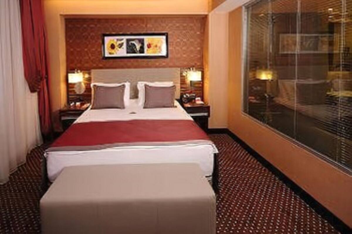 Habitación de hotel con una cama grande, dos mesitas de noche con lámparas, una ventana con persianas y una obra de arte colgada en la pared.