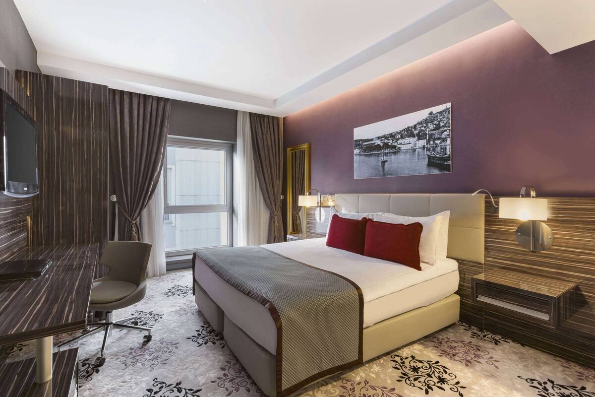 Habitación de hotel con cama, escritorio, silla y un ventanal con cortinas.