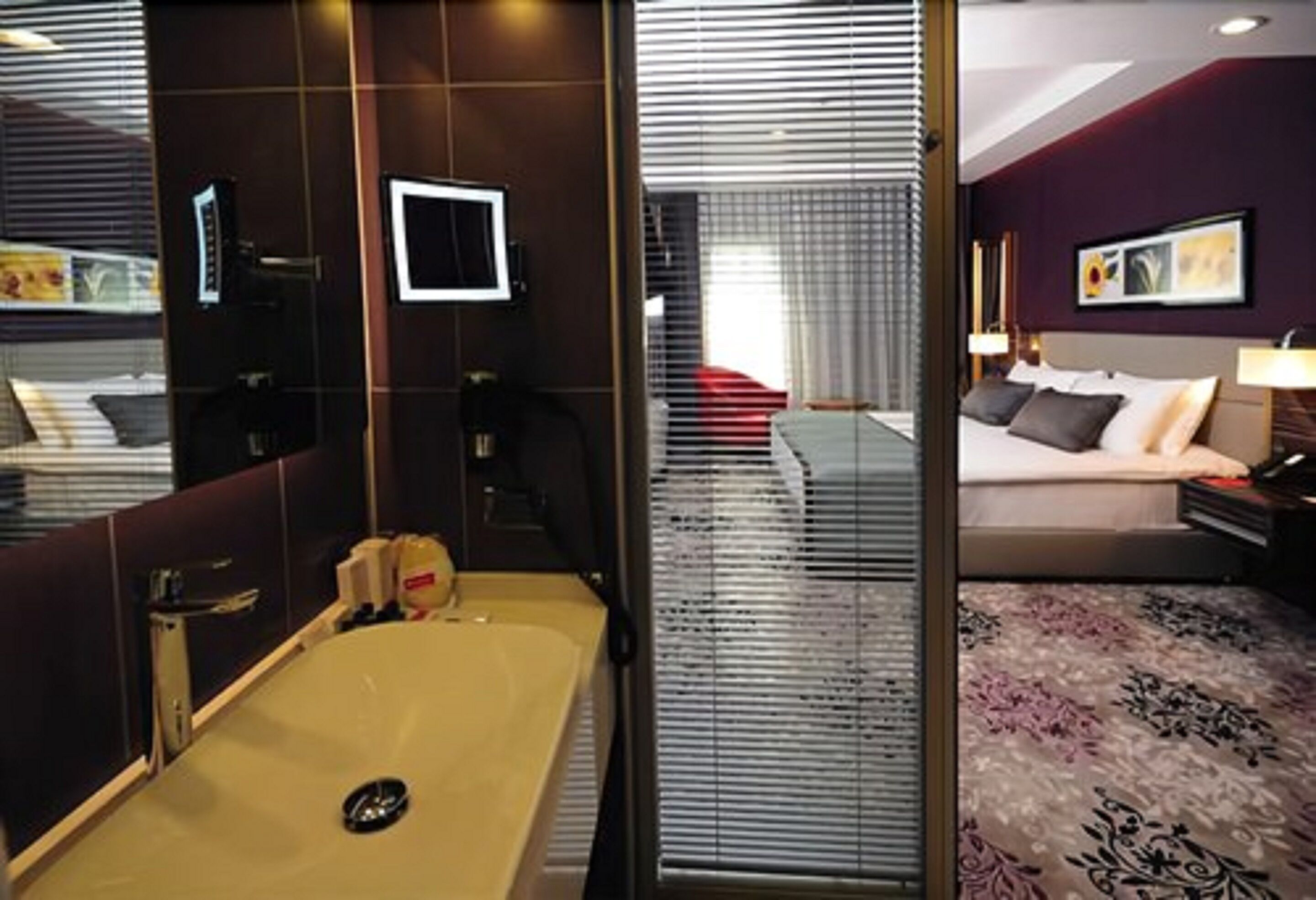 A modern hotel room with a large bed, a separate bathroom, and a view through a window with blinds.