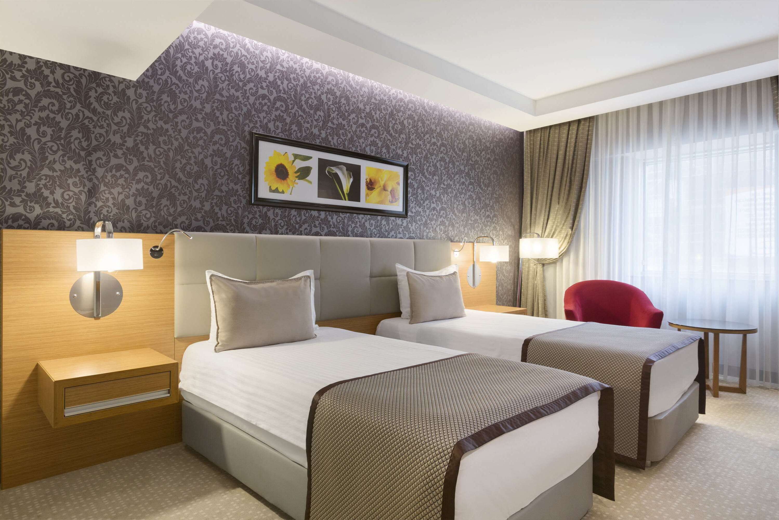 A hotel room with two beds, a red chair, a small table, and a wall with a floral pattern and a framed picture.