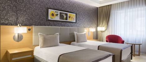 A hotel room with two beds, a red chair, a small table, and a wall with a floral pattern and a framed picture.