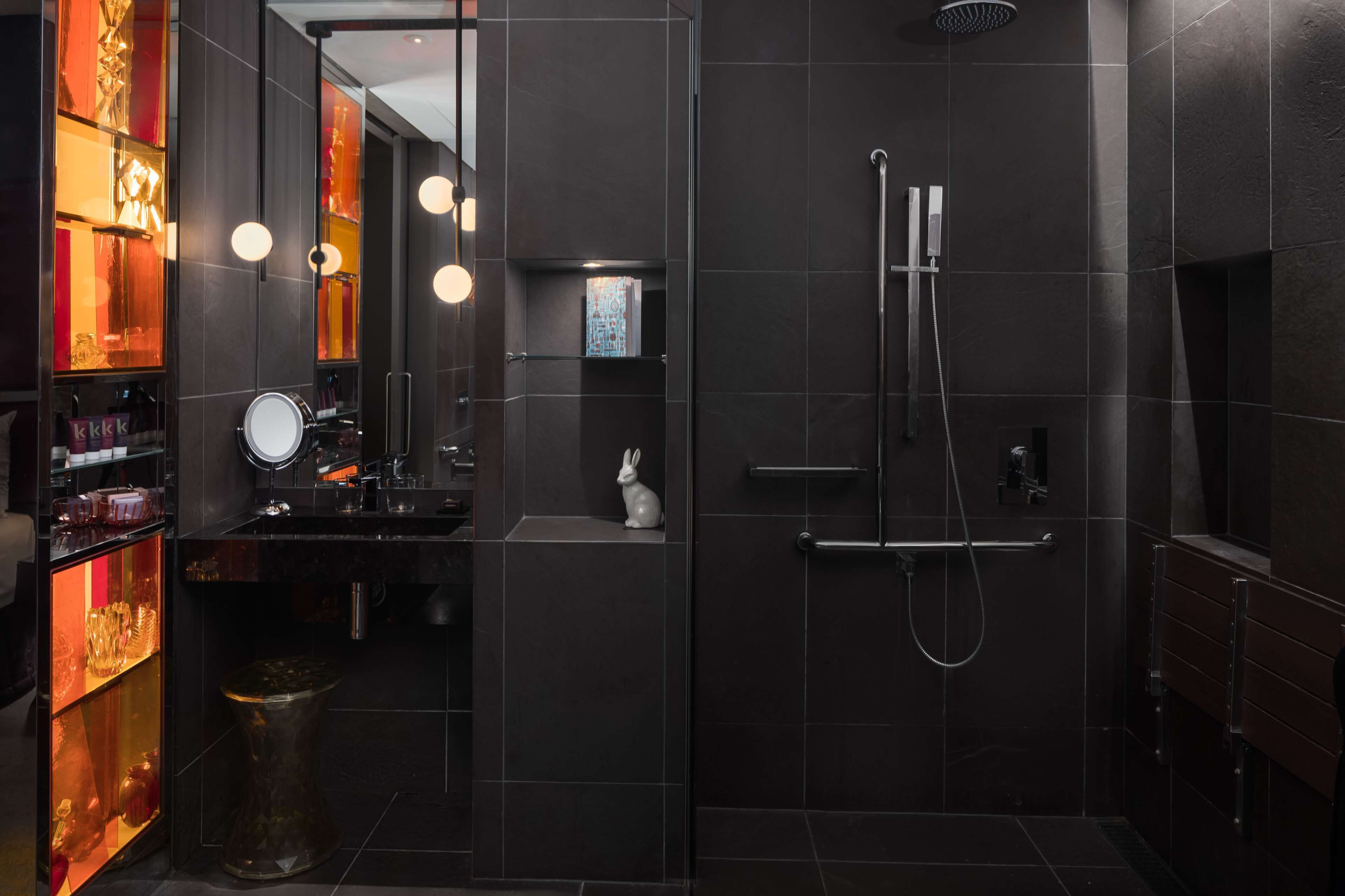 A modern bathroom with dark tiles, a shower, a vanity with a mirror, and a shelf with decorative items.