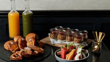 Une table dressée pour le petit-déjeuner avec des viennoiseries, des fruits, du granola et deux bouteilles de jus.