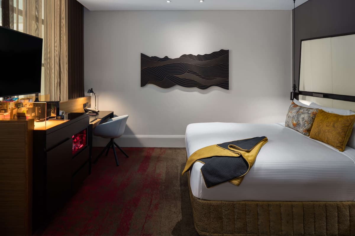 A modern hotel room with a bed, a desk, a chair, a flat-screen TV, and a wall art piece.