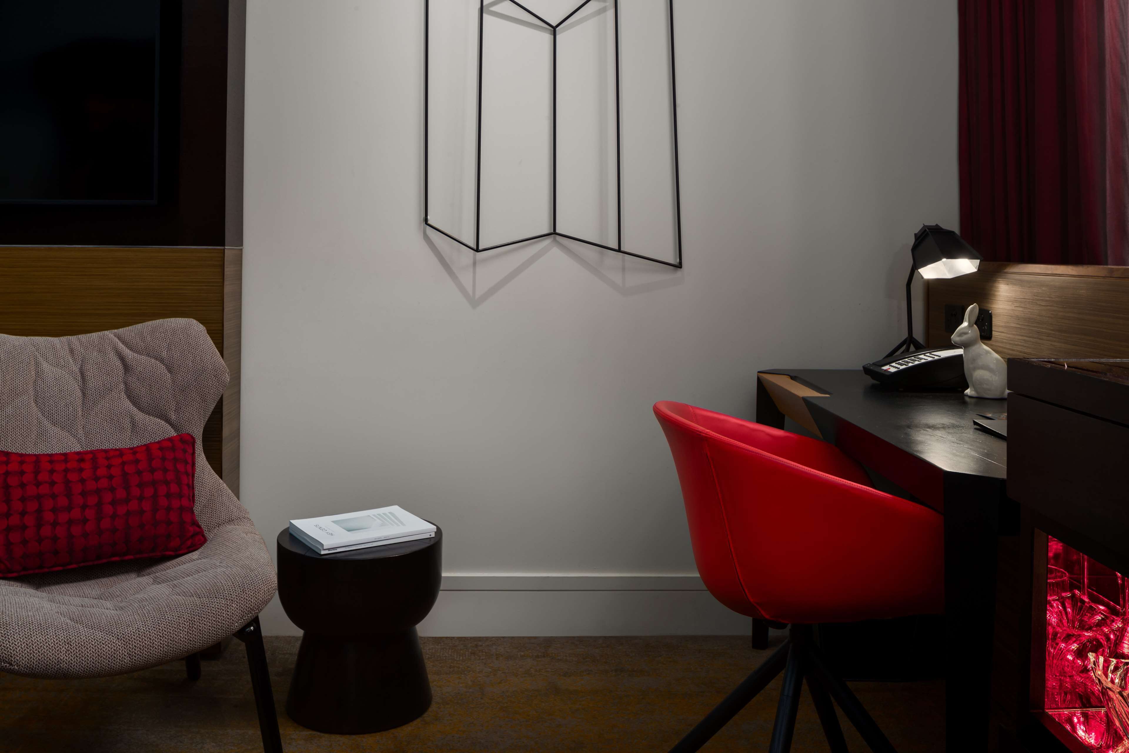 Une chambre moderne avec une tête de lit en bois, une chaise rouge, une petite table ronde et une lampe fixée au mur.