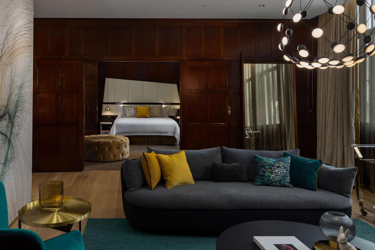 A modern hotel room with a dark grey sofa, a round coffee table, a tufted ottoman, and a large bed with a white headboard.