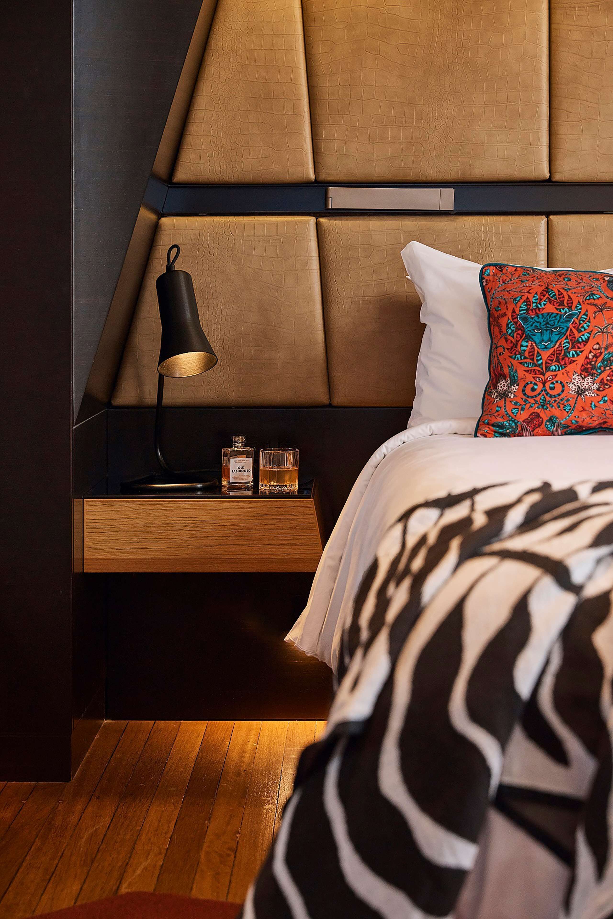 Un dormitorio moderno con cabecera de madera, una mesita de noche con lámpara, dos botellas de licor y un cojín estampado.