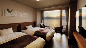Habitación de hotel con dos camas, un escritorio, una silla, un televisor y un ventanal con vista al atardecer sobre el agua.