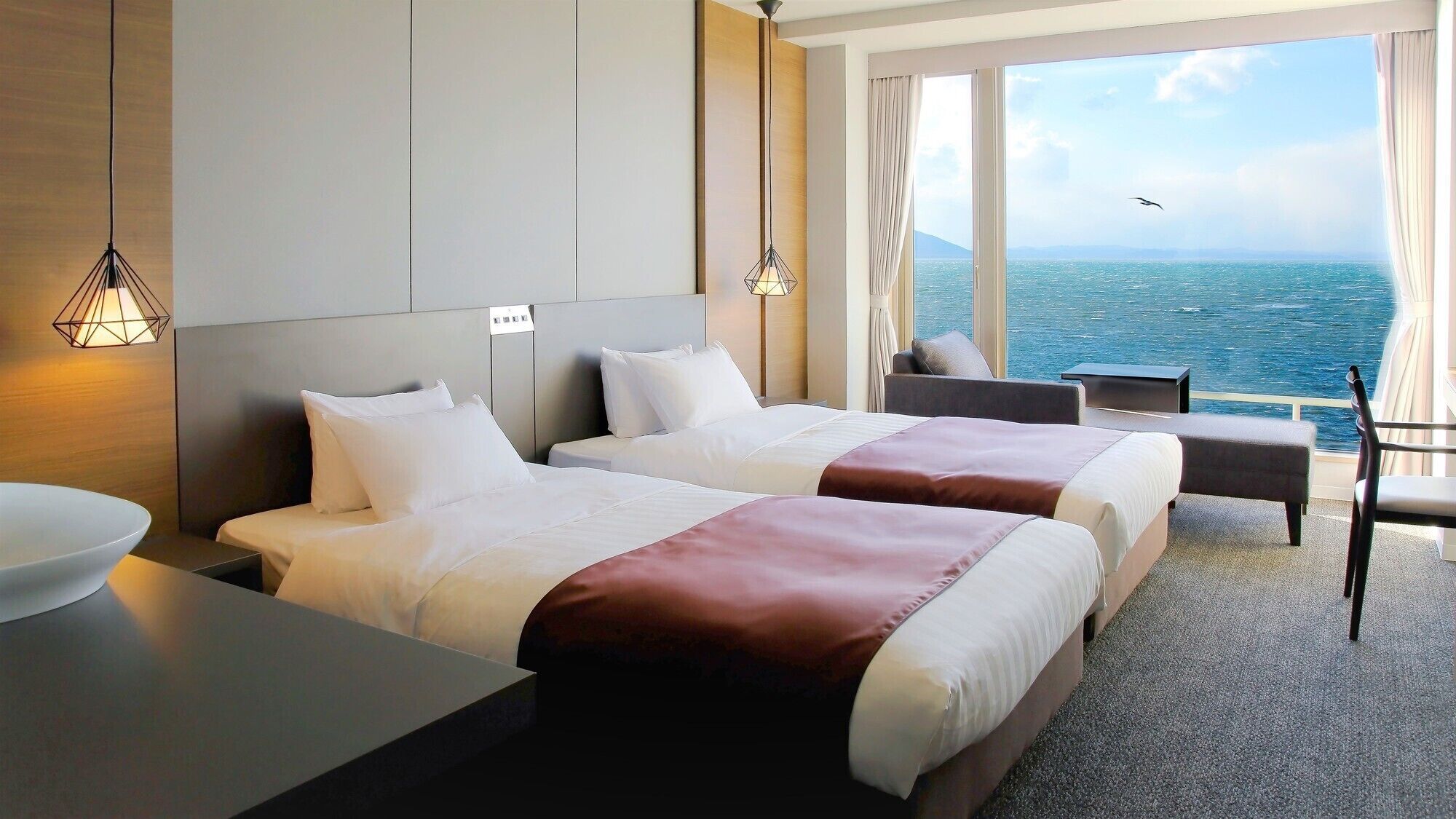 A hotel room with two beds, a view of the sea, a dining area, and a large window.