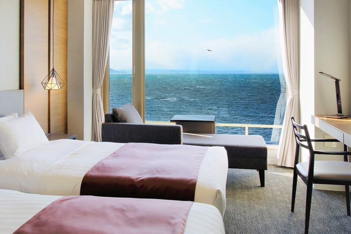 Habitación de hotel con dos camas, un escritorio, una silla y vista al mar.
