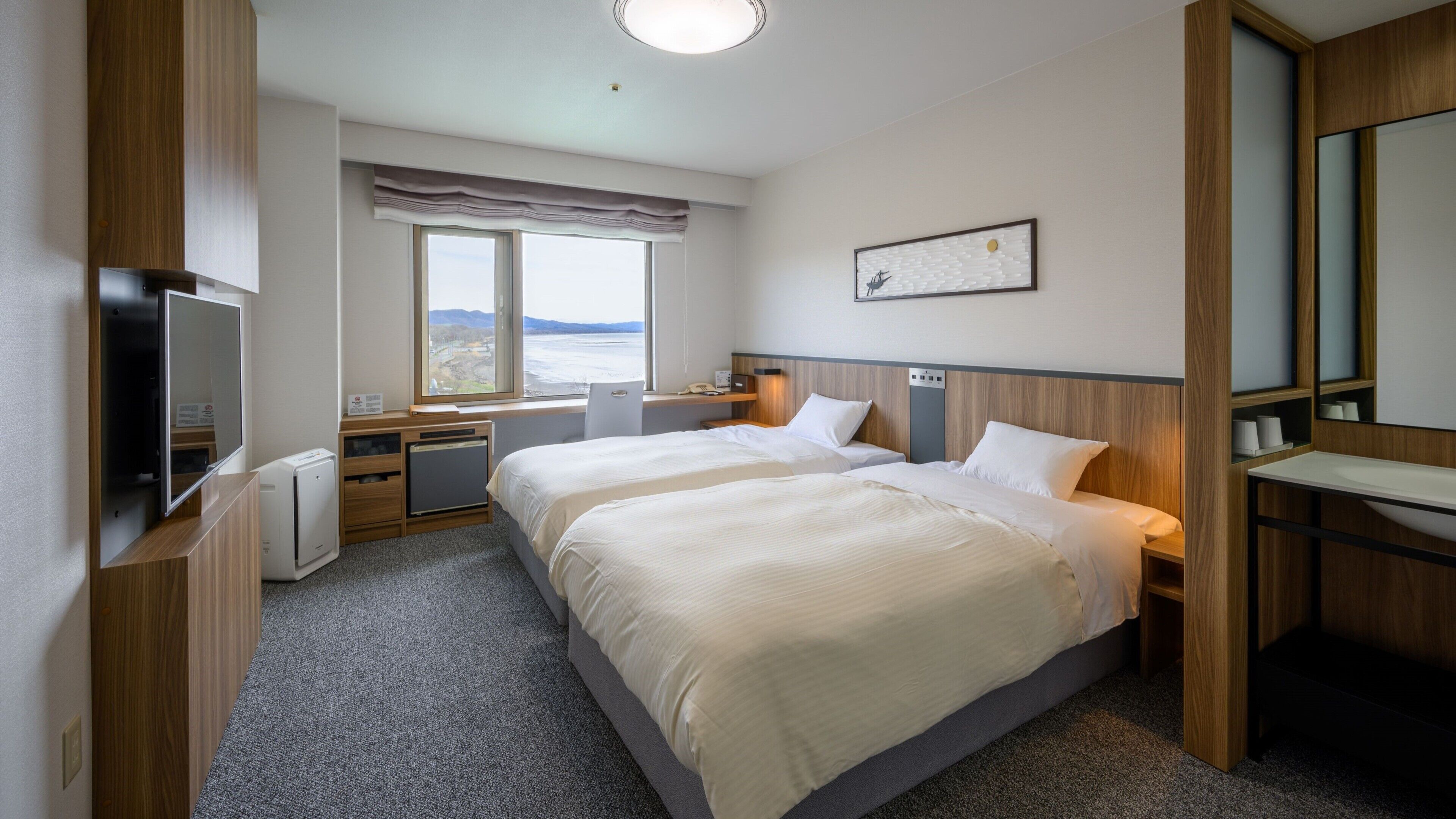 A hotel room with two beds, a desk, a TV, and a window with a view.