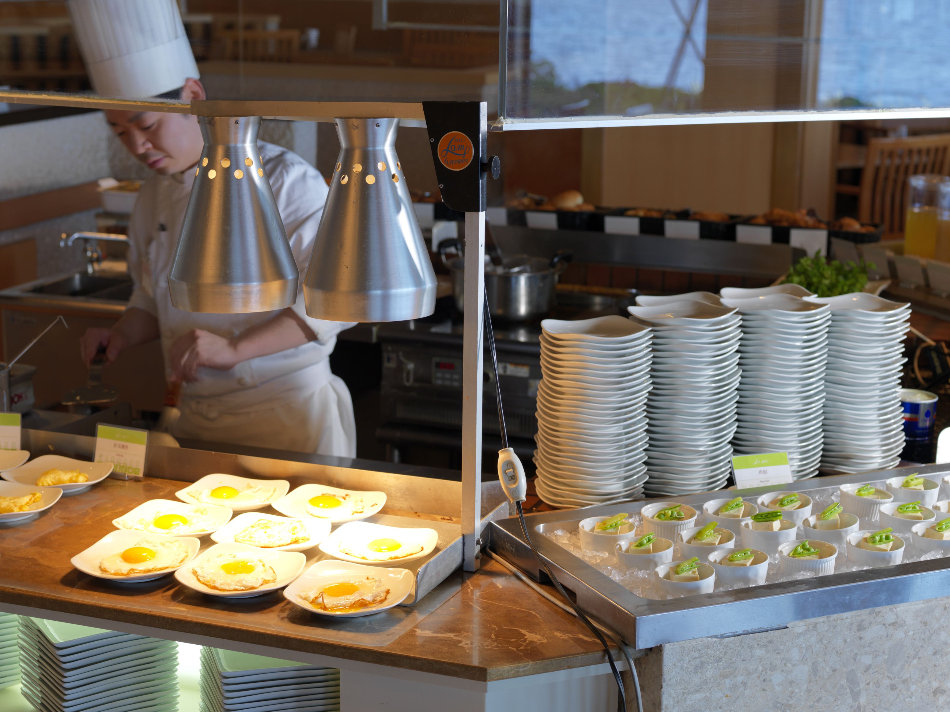 A buffet setup with plates of food, including fried eggs and desserts, and a chef in the background.
