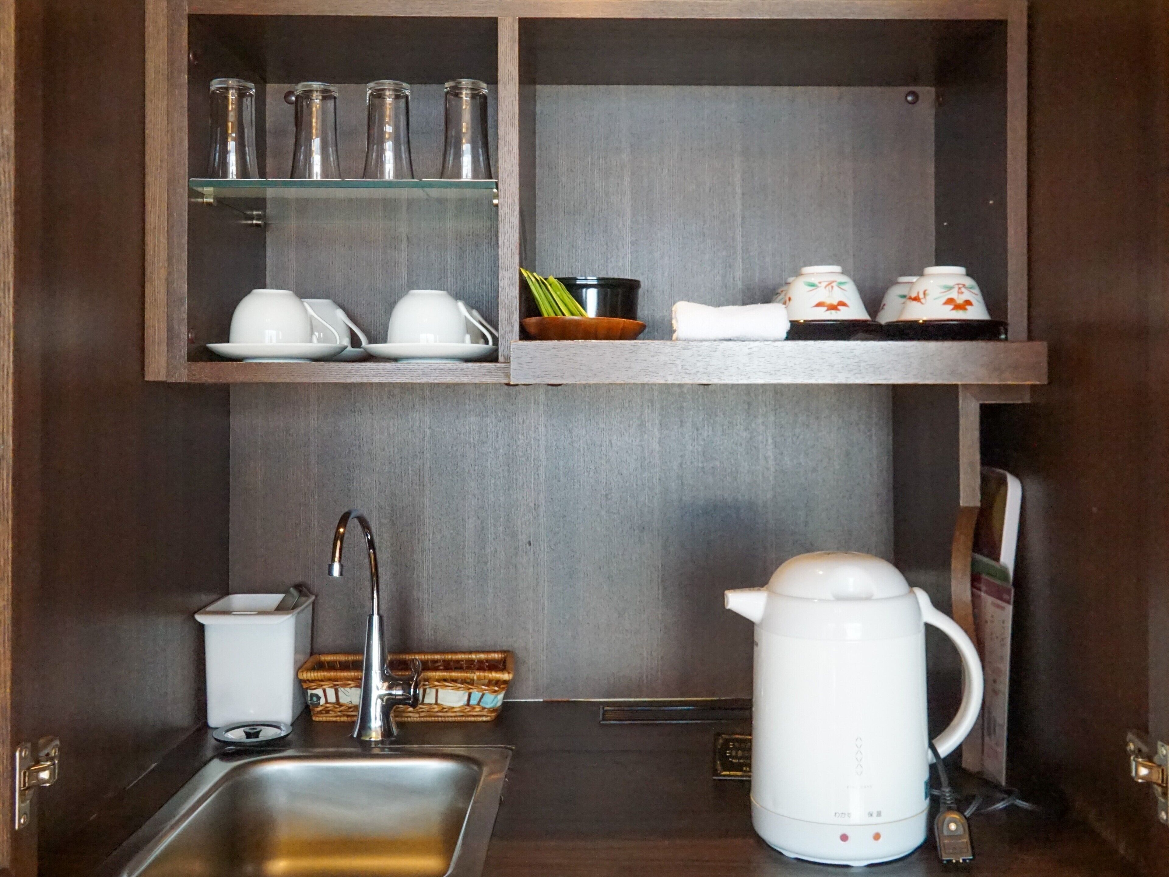 A kitchen with a stainless steel sink, a white electric kettle, and shelves above holding glass jars and ceramic cups.