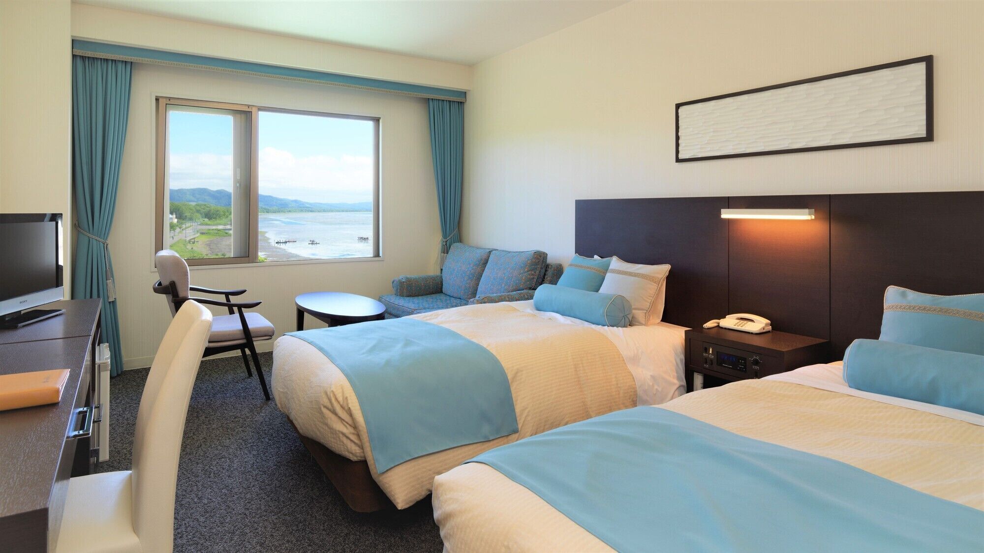 Habitación de hotel con dos camas, un escritorio con televisor, una silla, una mesita y vista al mar.