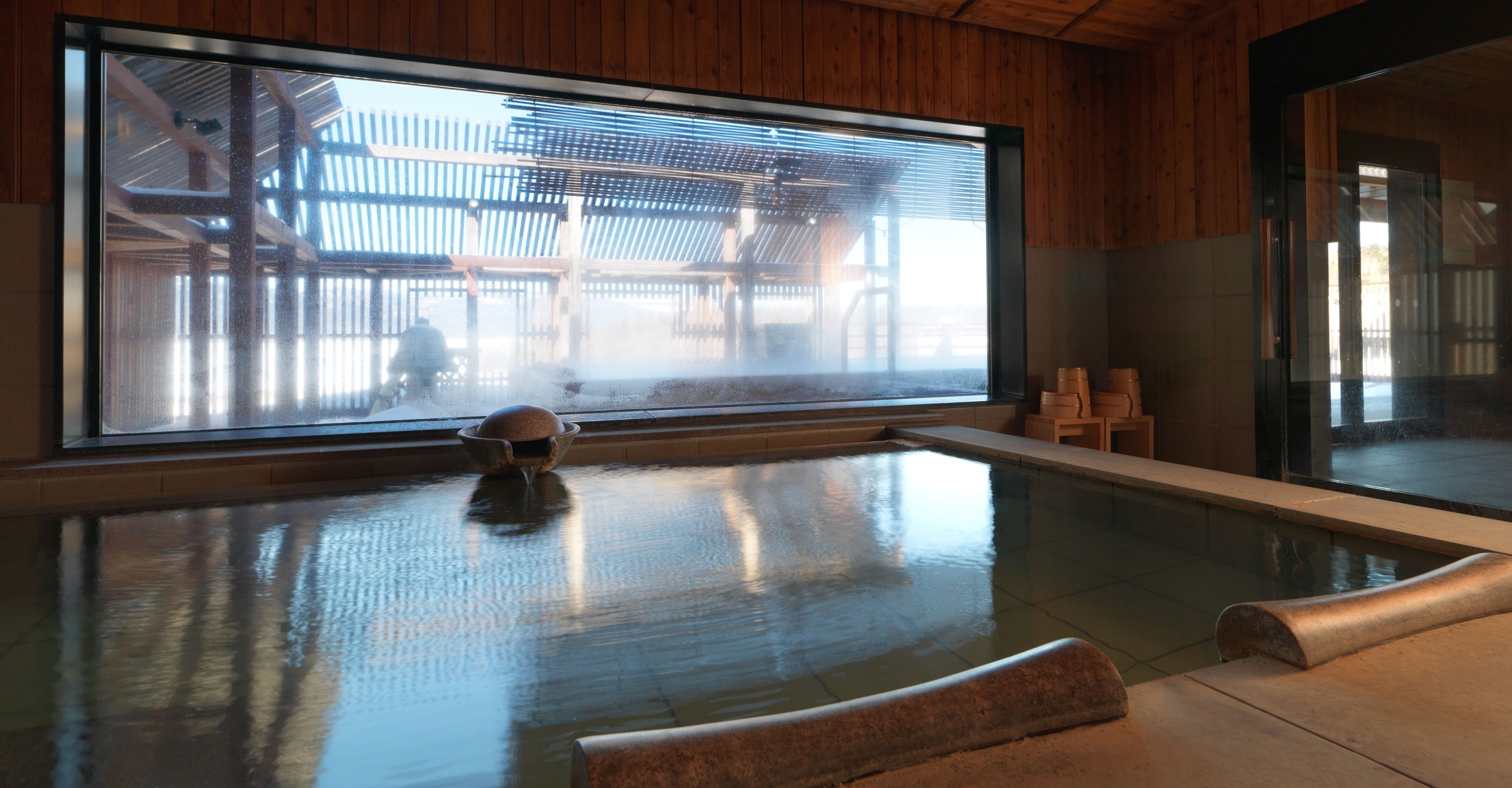 A modern indoor pool with a large window, wooden walls, and a stone seating area.