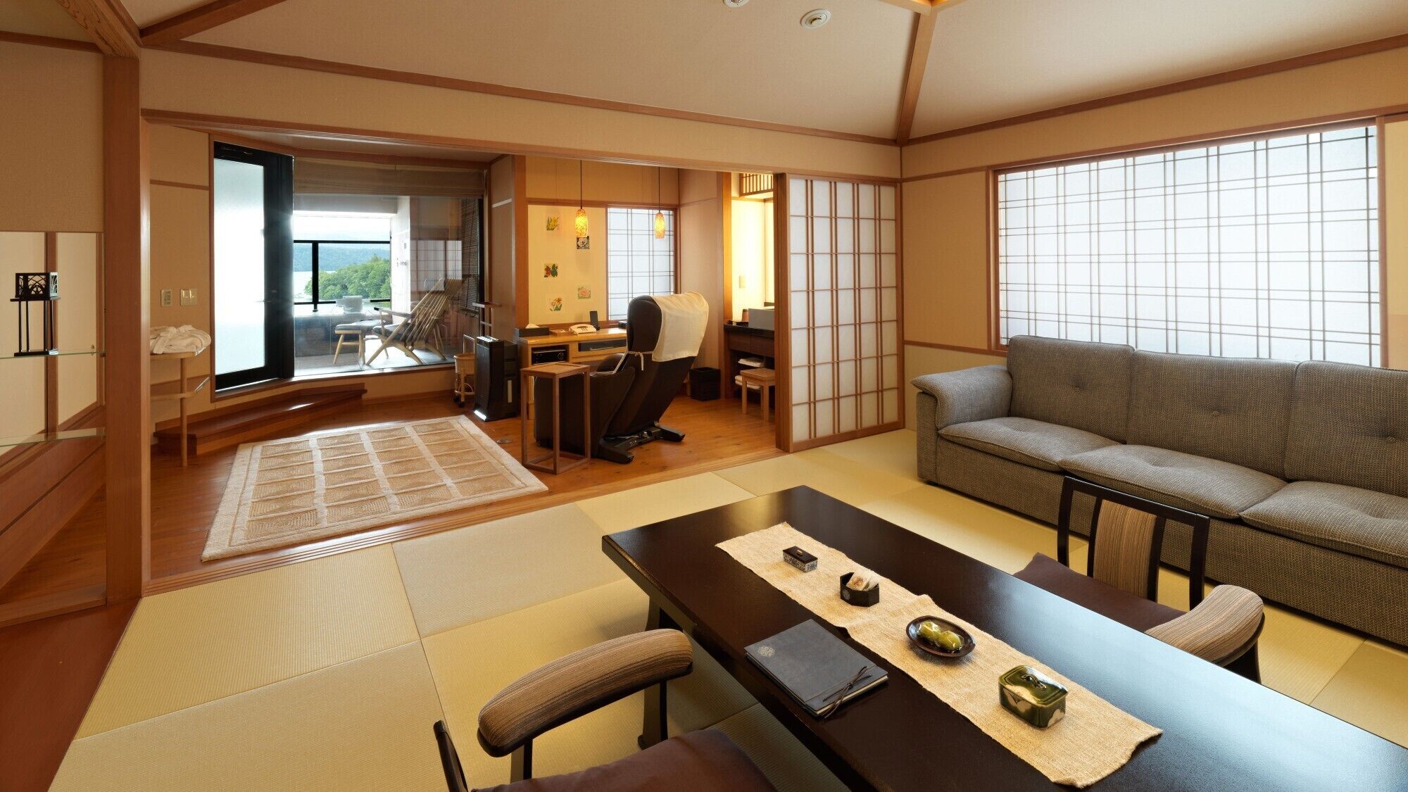 A traditional Japanese room with tatami flooring, a tatami mat in the center, a sofa, a desk with a chair, and a sliding door leading to a balcony.