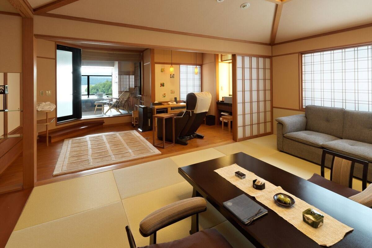 A traditional Japanese room with tatami flooring, a tatami mat in the center, a sofa, a desk with a chair, and a sliding door leading to a balcony.