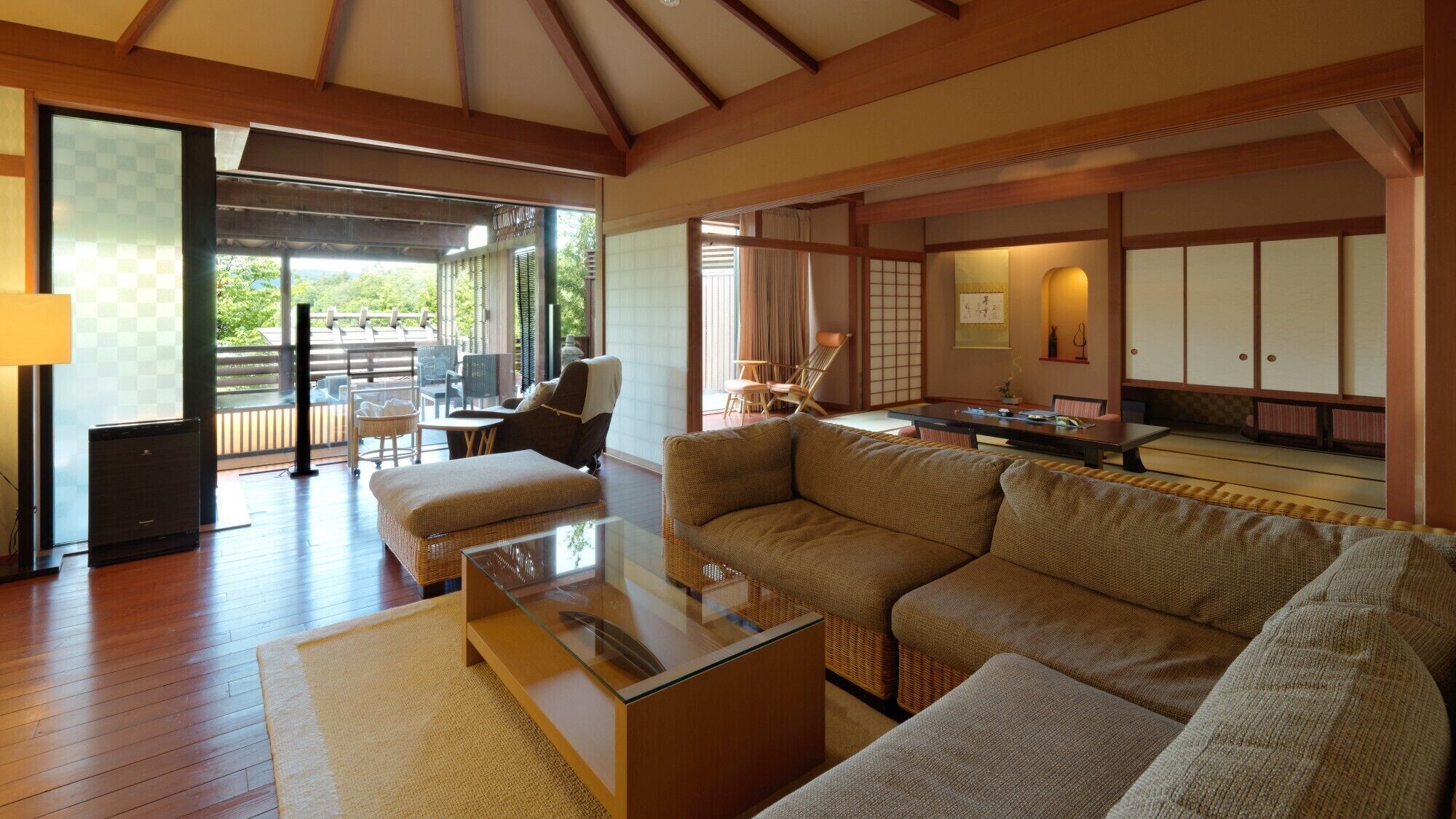 A spacious living area with a large sofa, a glass coffee table, and a dining table. There is a sliding glass door leading to an outdoor deck.