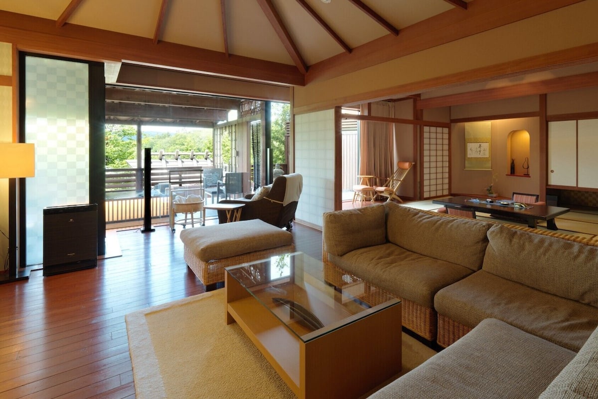 A spacious living area with a large sofa, a glass coffee table, and a dining table. There is a sliding glass door leading to an outdoor deck.