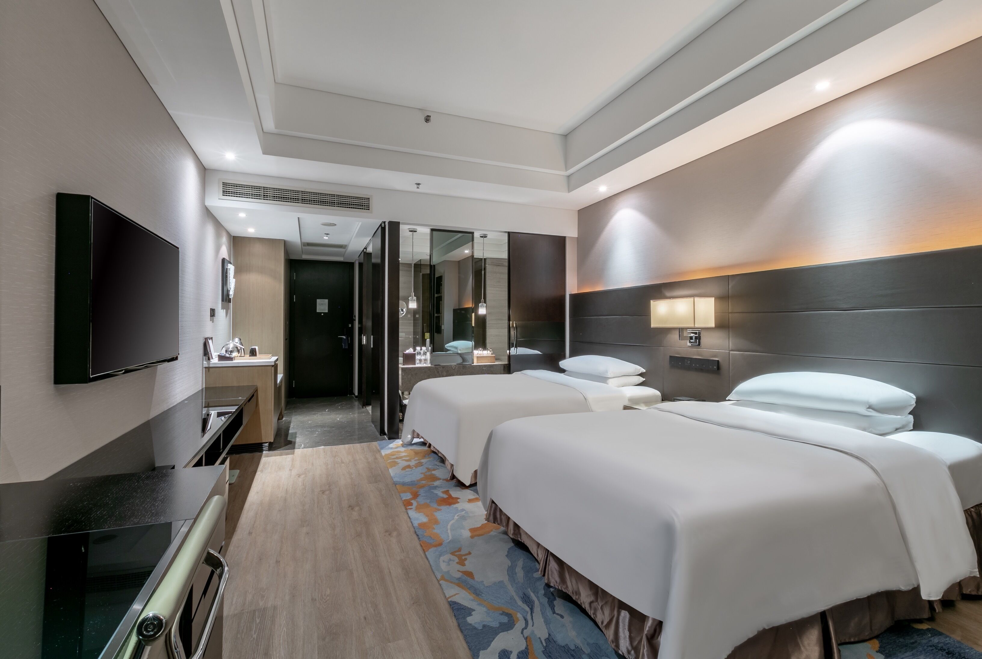 A hotel room with two beds, a flat-screen TV, a desk, and a bathroom visible through a glass door.