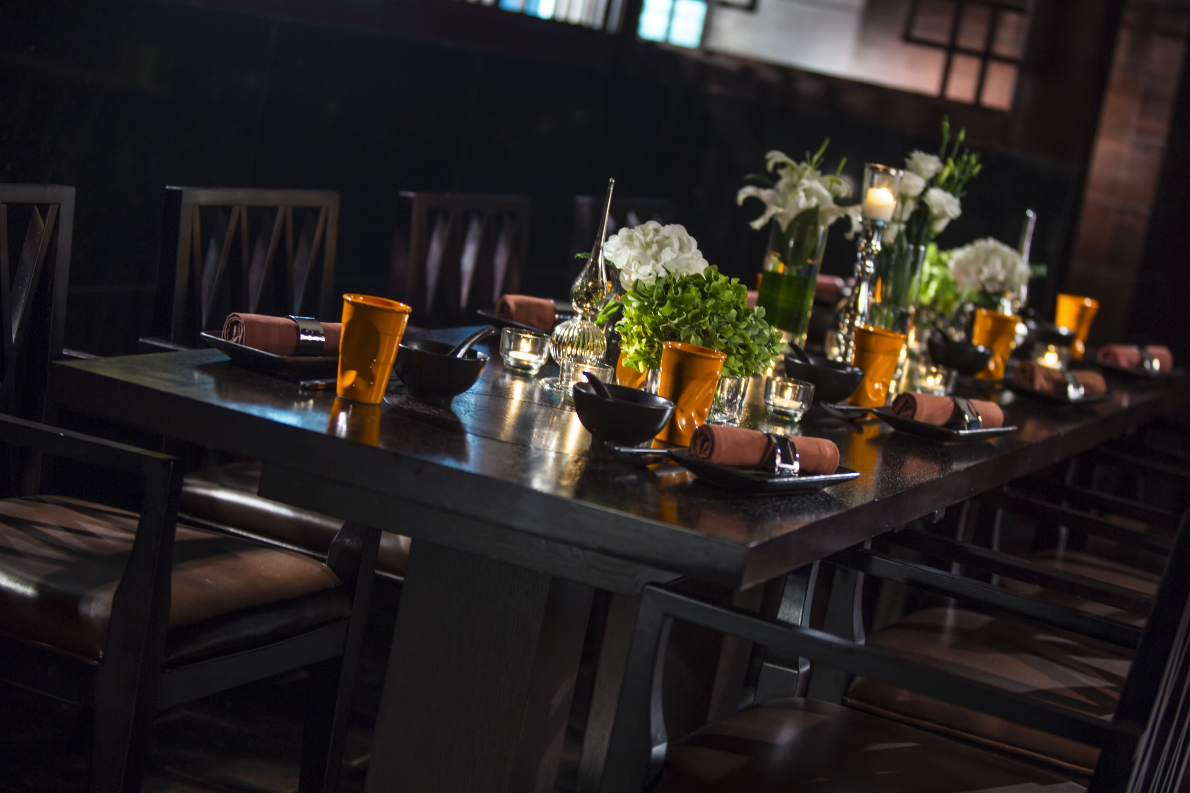 Una larga mesa de comedor adornada con cristalería naranja, flores blancas y velas, rodeada de sillas de madera oscura.