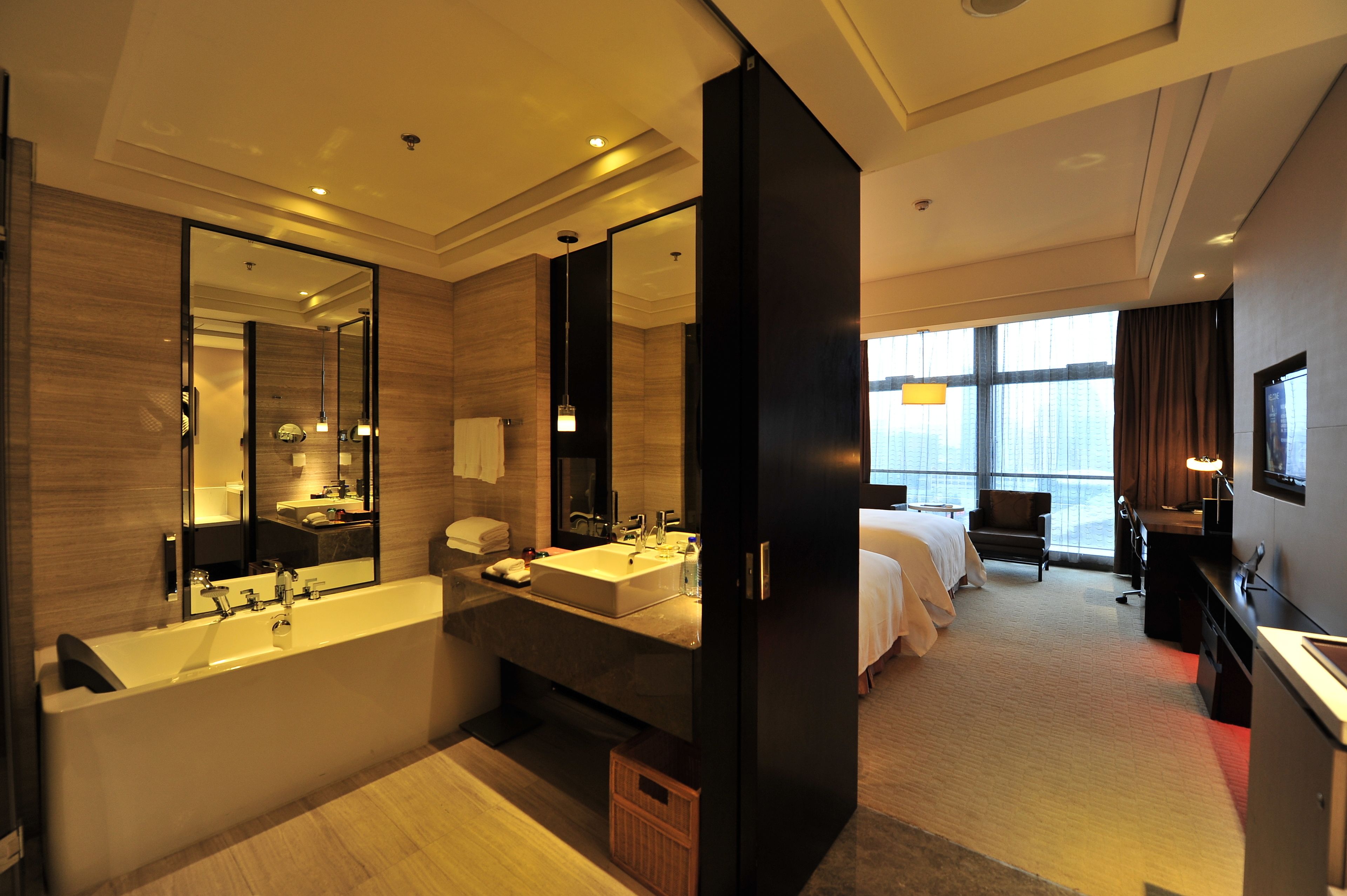 A modern hotel bathroom with a large bathtub, a sink, and a mirror. There is a bed with a desk and a chair in the adjacent room.