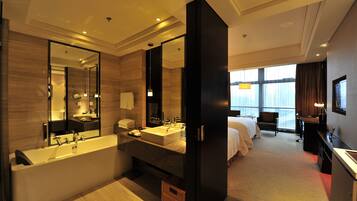 A modern hotel bathroom with a large bathtub, a sink, and a mirror. There is a bed with a desk and a chair in the adjacent room.