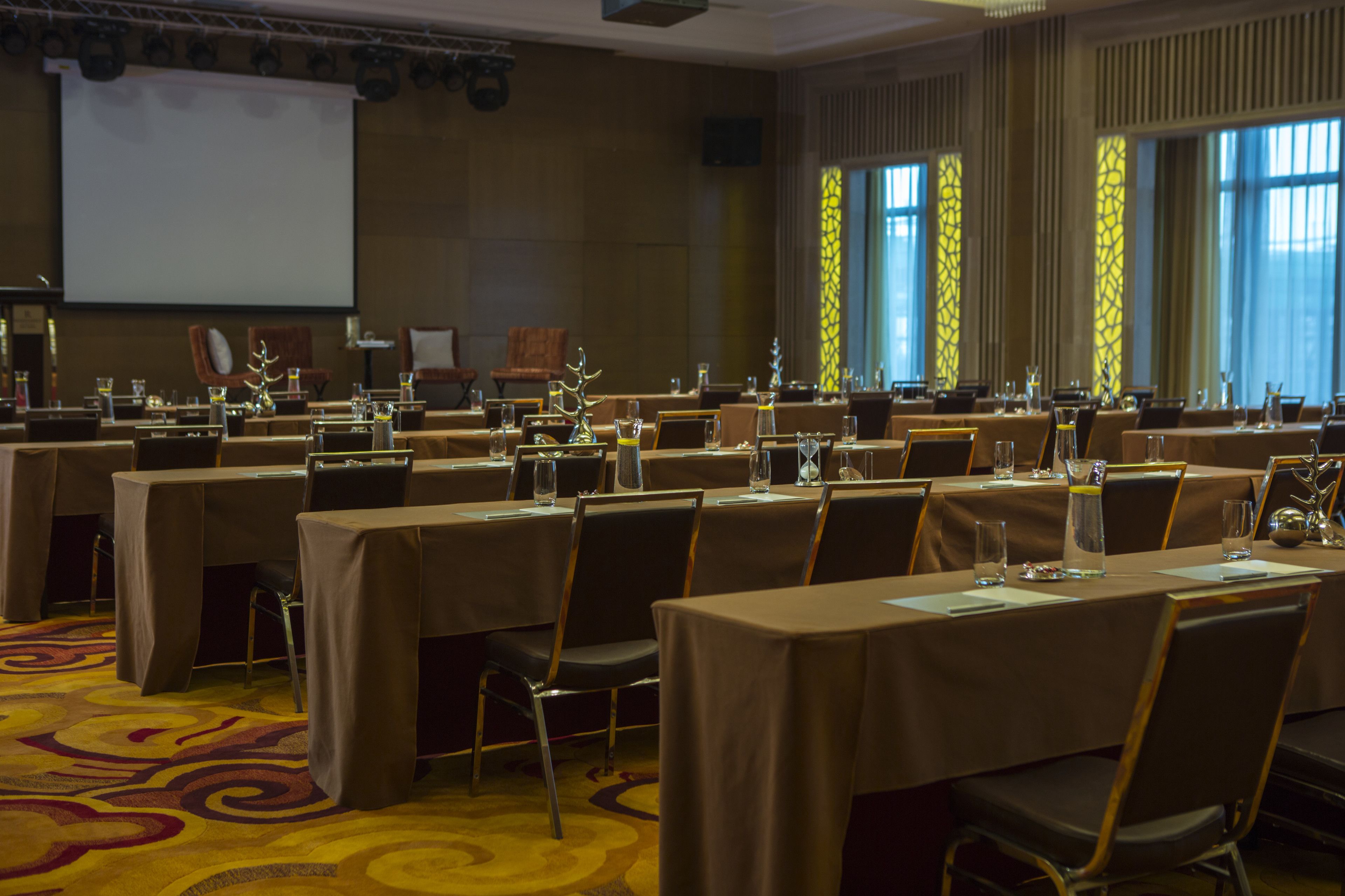 A conference room with multiple tables and chairs, a projector screen, and large windows with patterned curtains.