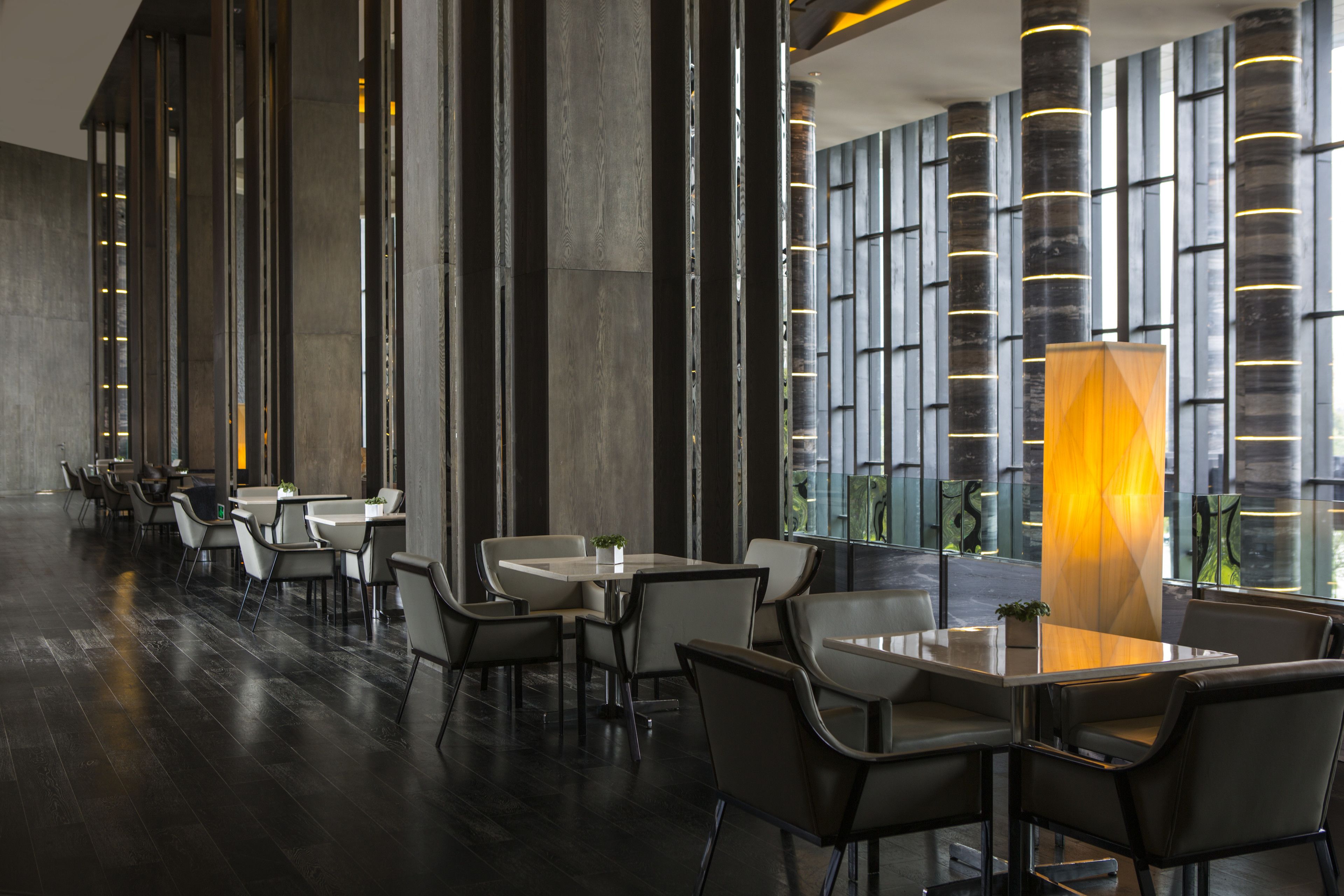 A modern hotel lobby with a long row of tables and chairs, large windows, and a distinctive illuminated column.