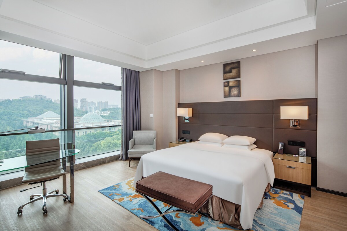 A modern hotel room with a large bed, a desk, a chair, and a view of a cityscape through large windows.