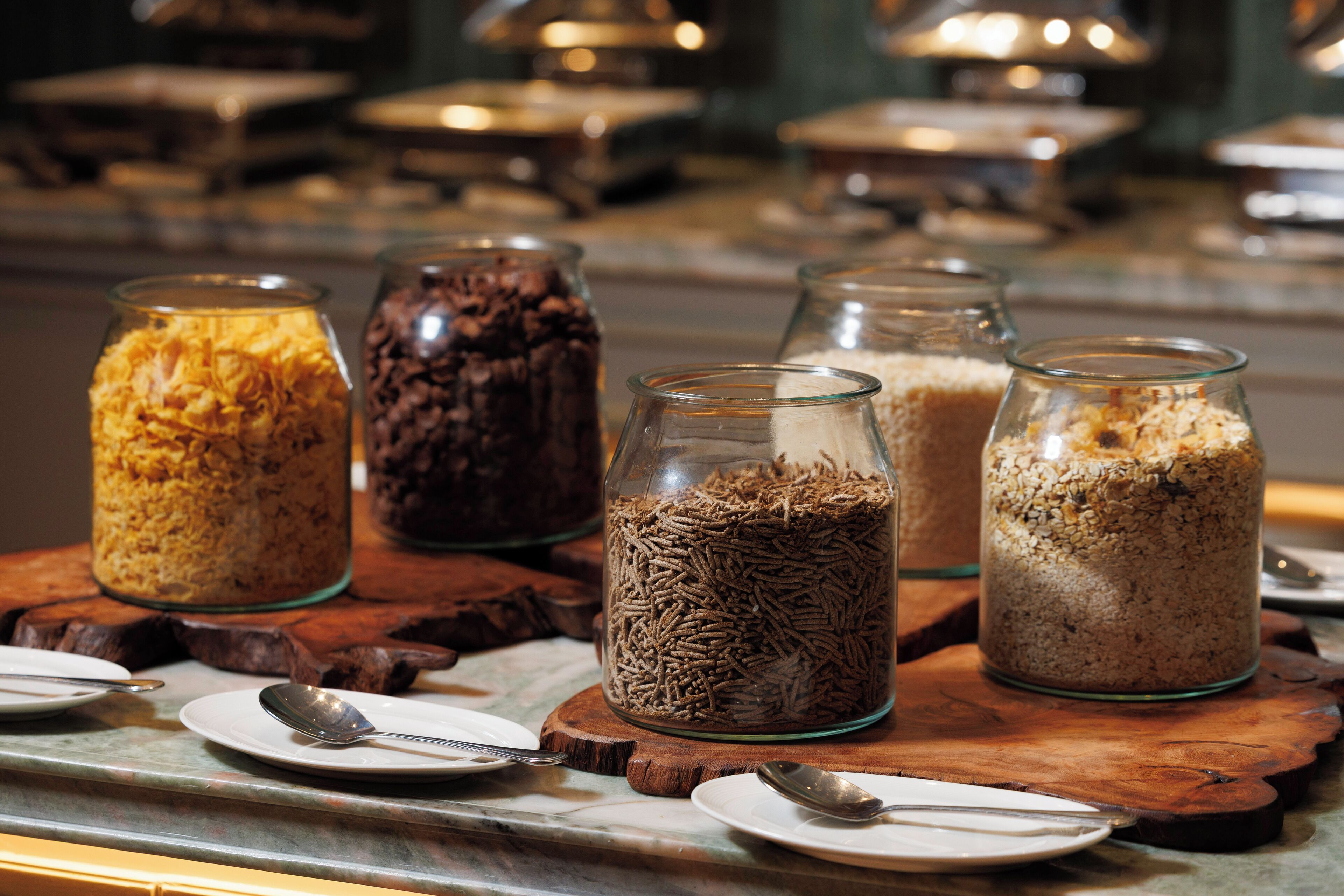 Uma mesa de buffet com diversos alimentos em recipientes de vidro, acompanhados de pratos e colheres.