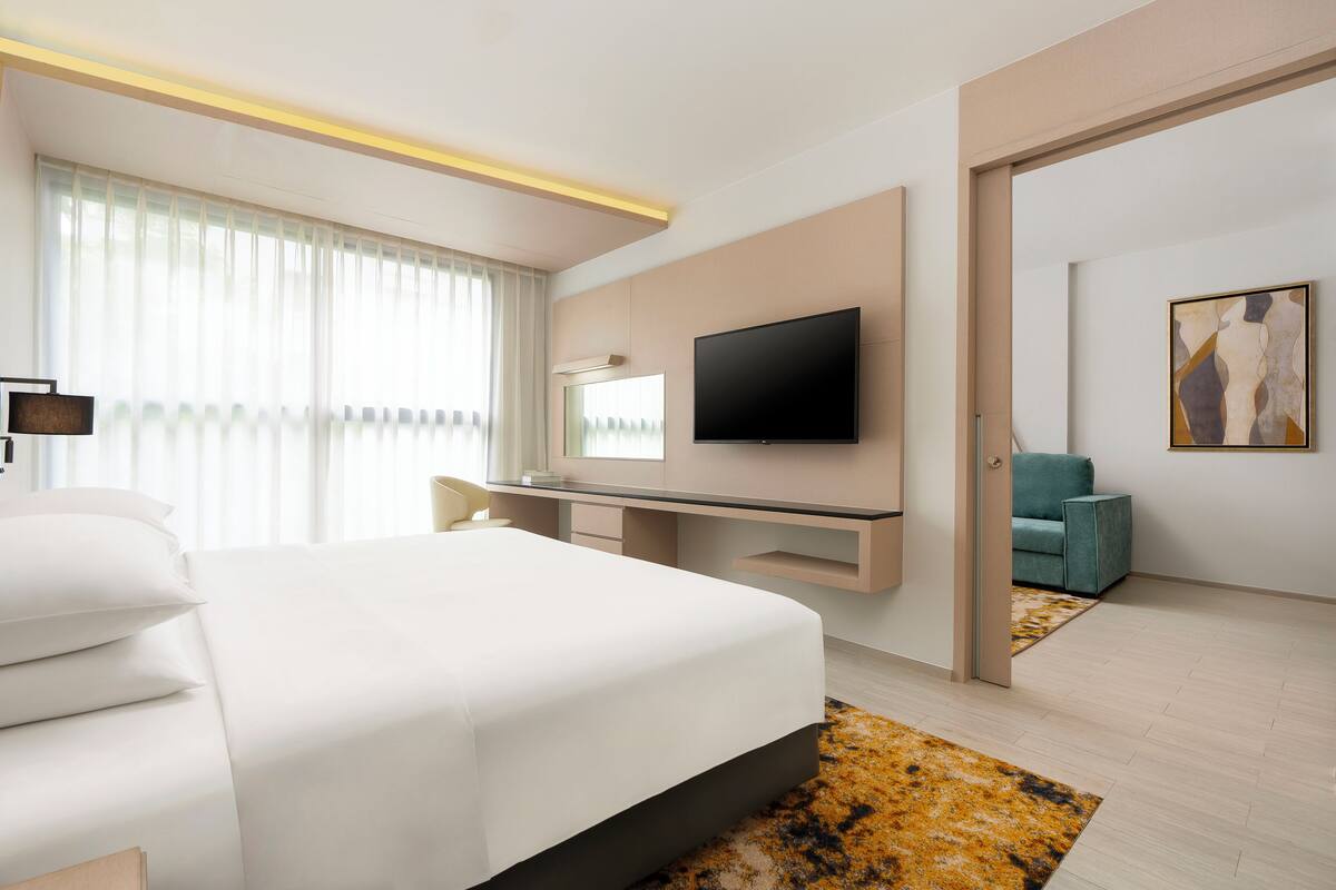 Una habitación de hotel moderna con una cama grande, un escritorio con un televisor de pantalla plana, una zona de estar con una silla color turquesa y un cuadro en la pared.