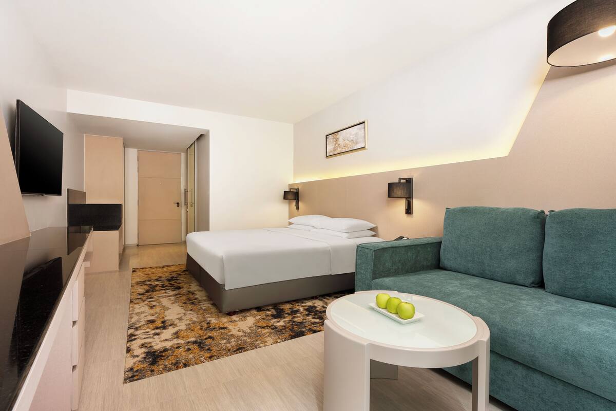 Una habitación de hotel moderna con una cama, un sofá verde, una mesita con manzanas, un televisor de pantalla plana y una lámpara fijada a la pared.