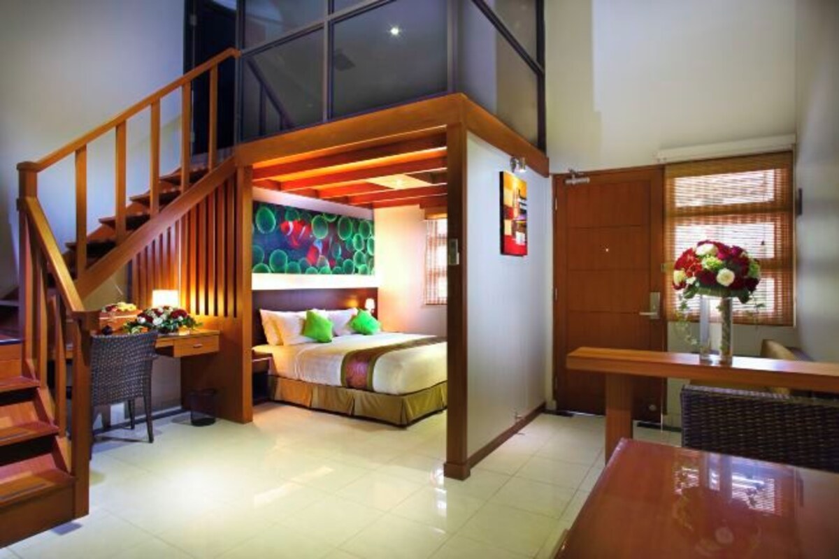 Una habitación de hotel compacta con cama en altillo, escalera de madera, zona de comedor y un ventanal grande con persianas.