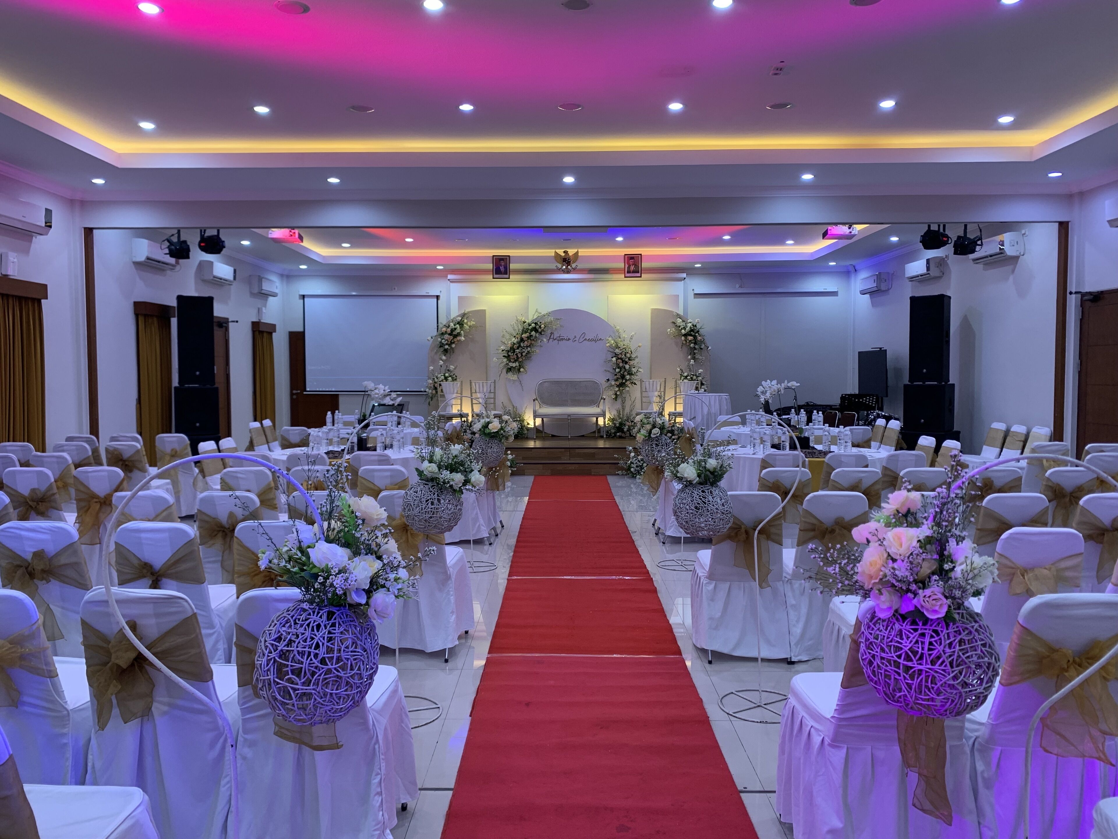 Un salón de bodas con hileras de sillas blancas adornadas con cintas doradas, una alfombra roja en el pasillo y decoraciones florales.