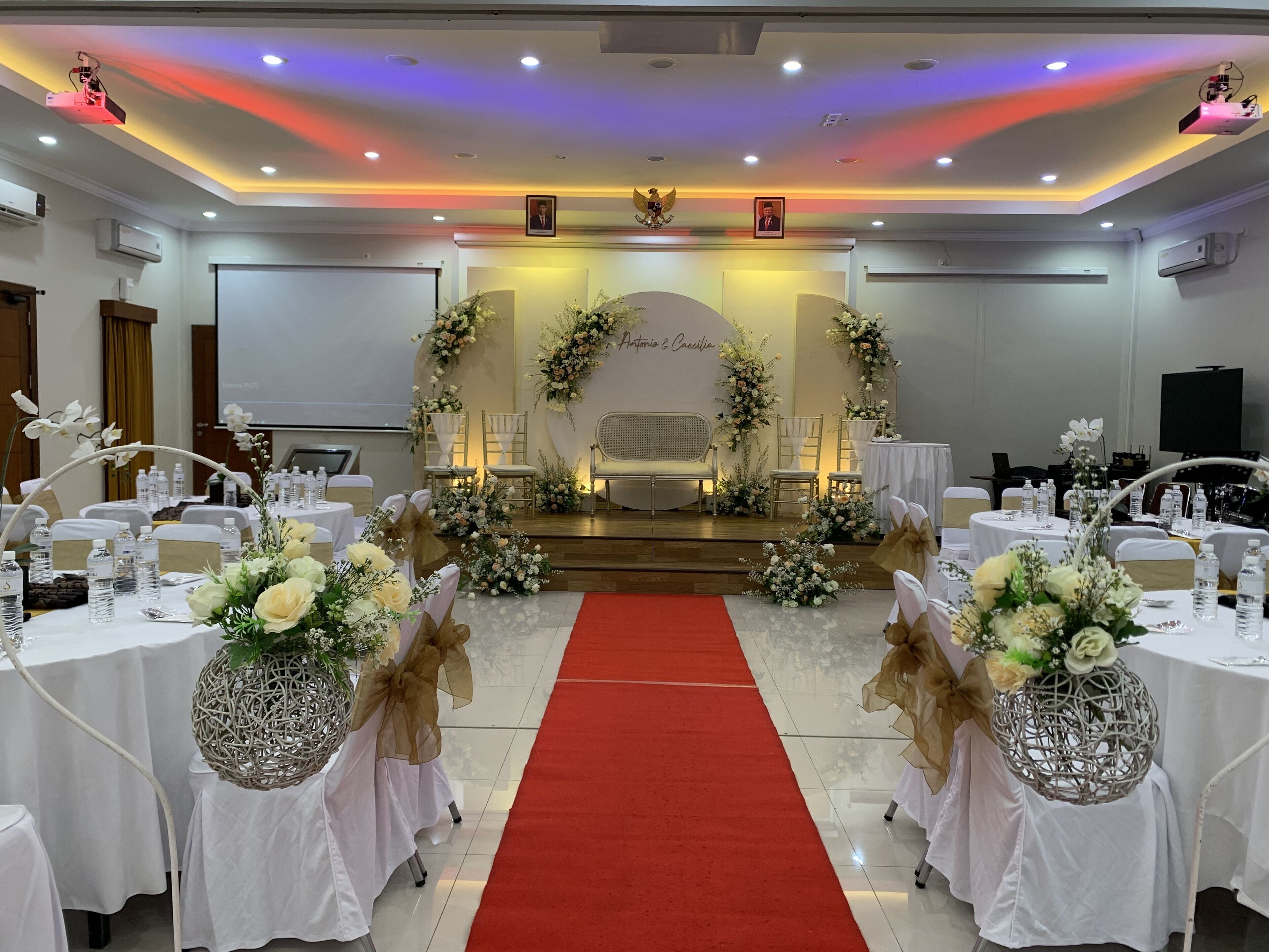 A conference room with a stage, white tables, chairs, and floral decorations.