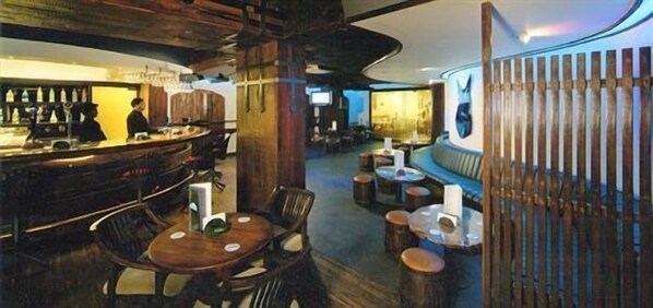 Una zona de bar con mobiliario de madera y una persona de pie junto a la barra. Hay taburetes y una barra alta con botellas.