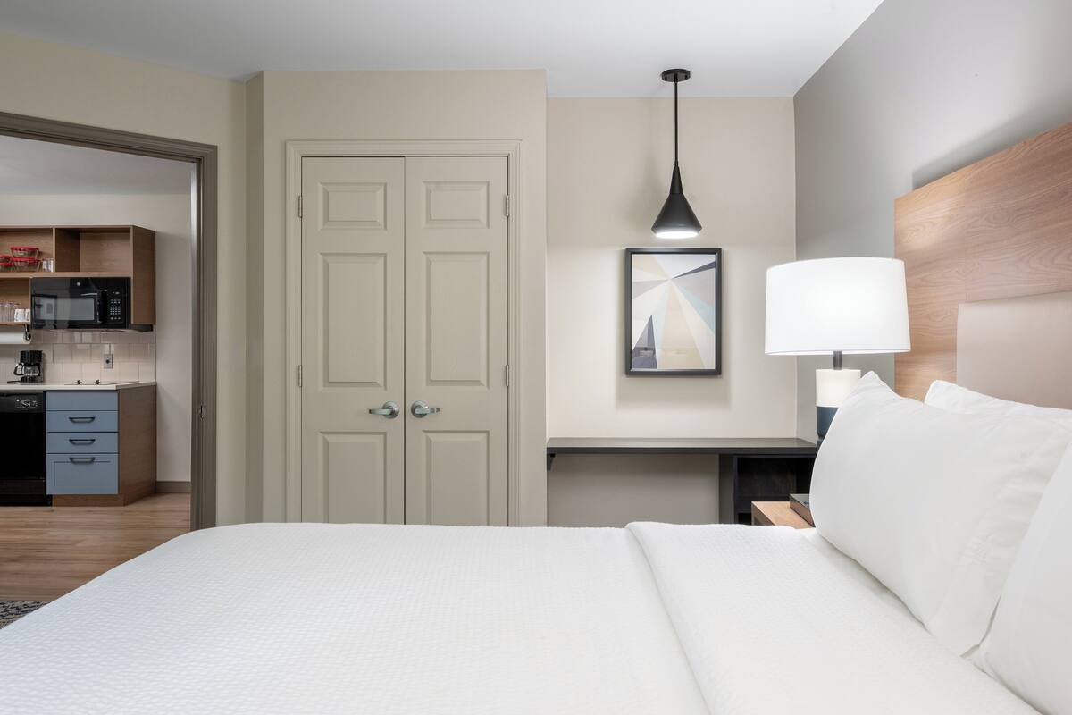 Une chambre d’hôtel avec un lit, un bureau avec une lampe, un four à micro-ondes et une porte donnant sur une petite cuisine.