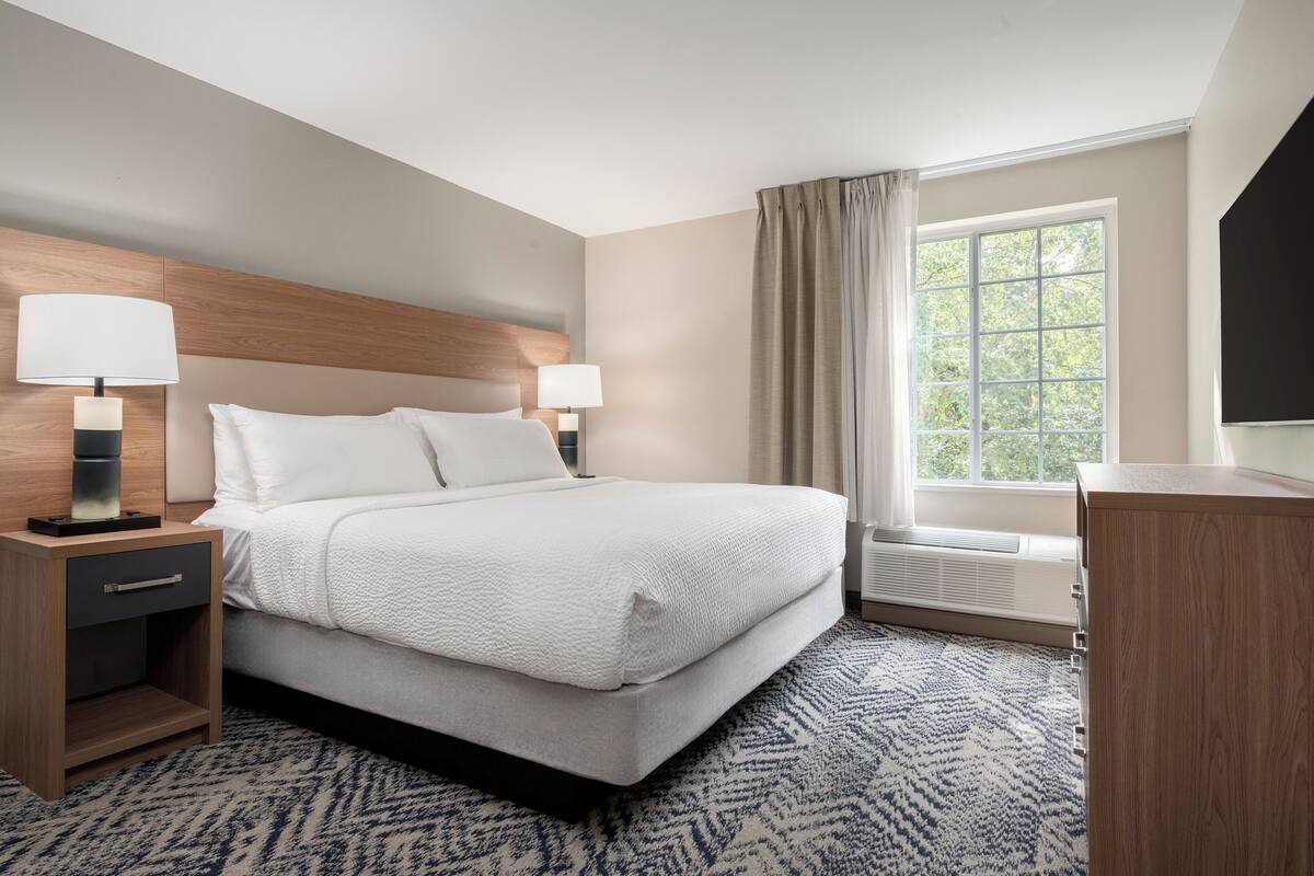 Une chambre d’hôtel comprenant un lit, deux tables de chevet avec des lampes, une fenêtre avec des rideaux et un téléviseur à écran plat posé sur un meuble en bois.