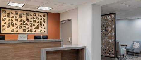 Een receptiezone met een houten receptiebalie, een muur met geometrische patronen, en een zithoek met een stoel en een klein tafeltje.