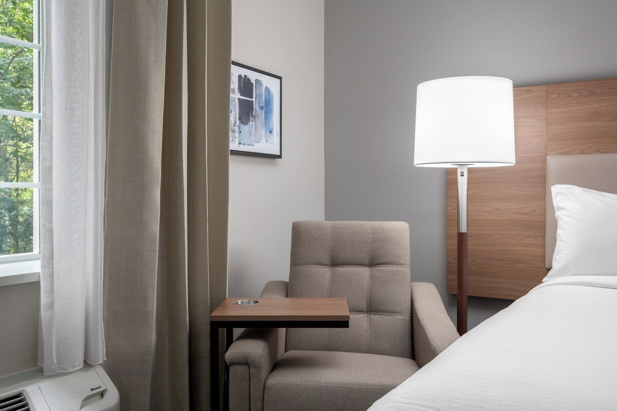 Ein Hotelzimmer mit Bett, Stuhl, kleinem Tisch, Lampe und einem Fenster mit Vorhängen.