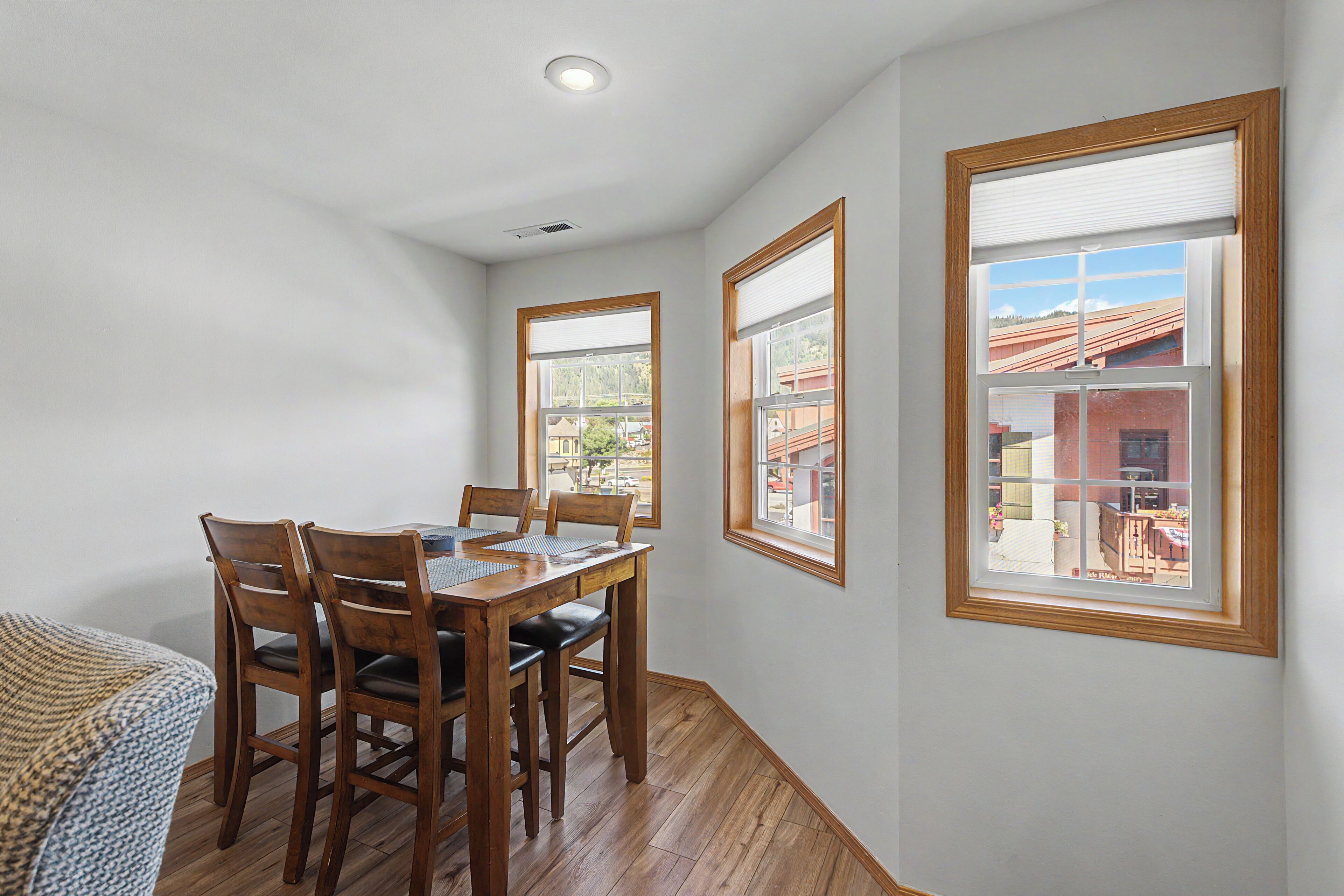 Un comedor con mesa y sillas de madera, ventanas amplias con persianas y vistas a edificios.