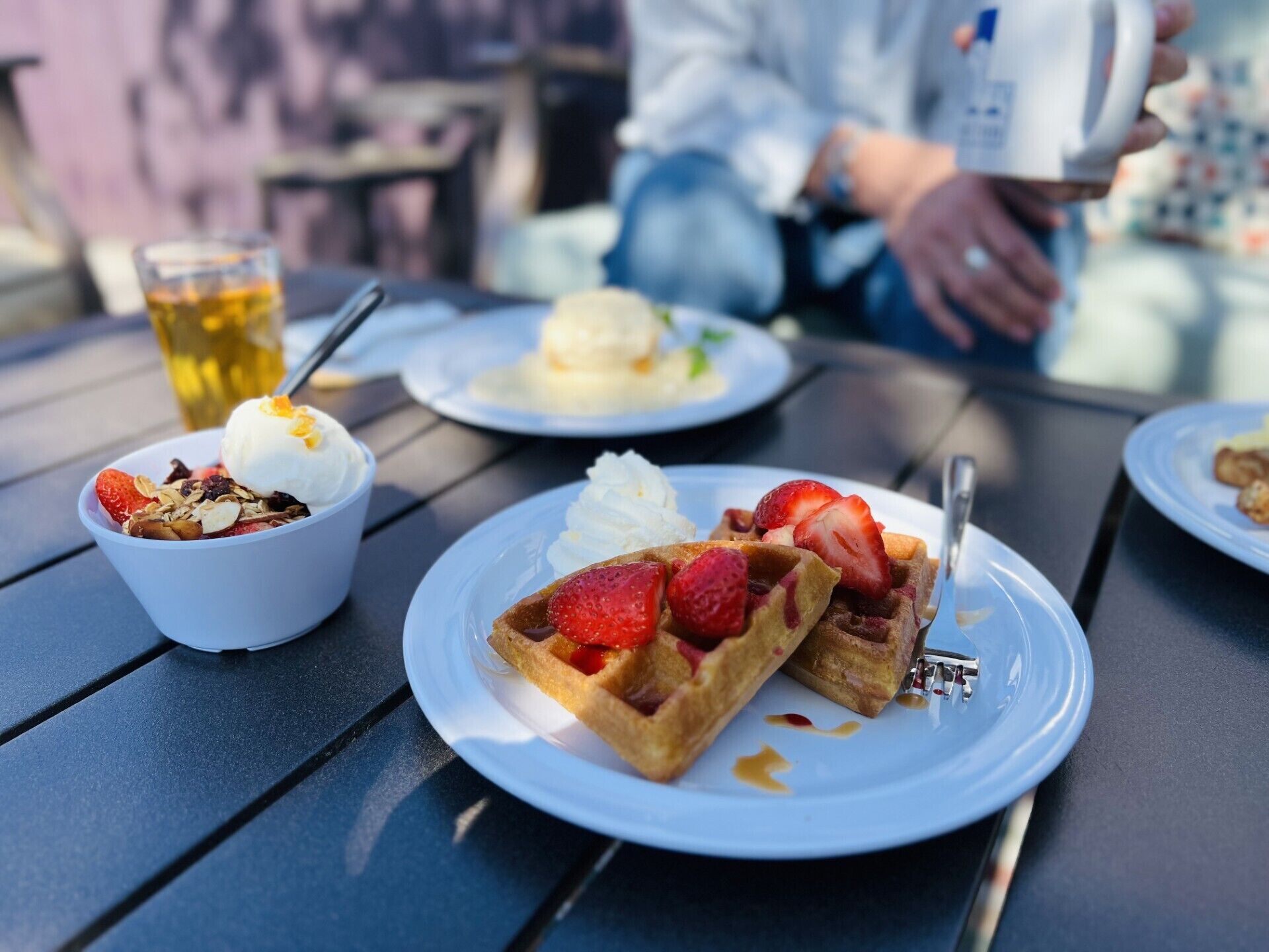 Una mesa con un plato de gofres cubiertos de fresas y crema batida, un tazón de yogur con fruta y un vaso de una bebida amarilla.