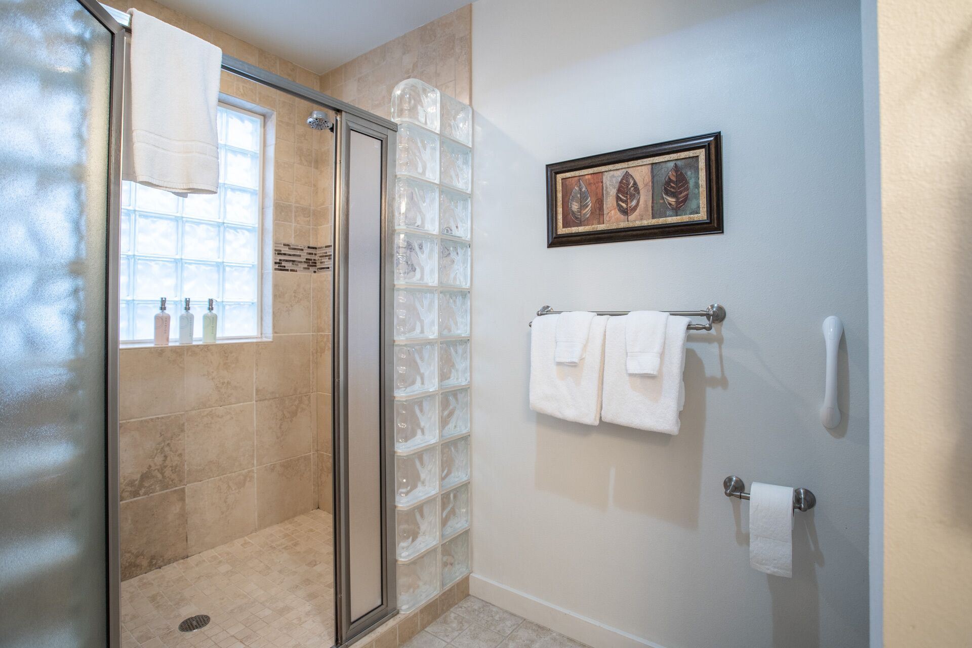 Une salle de bain avec une porte de douche en verre, un espace de douche carrelé, une fenêtre ornée de bouteilles, une œuvre d'art encadrée et un porte-serviettes avec des serviettes.